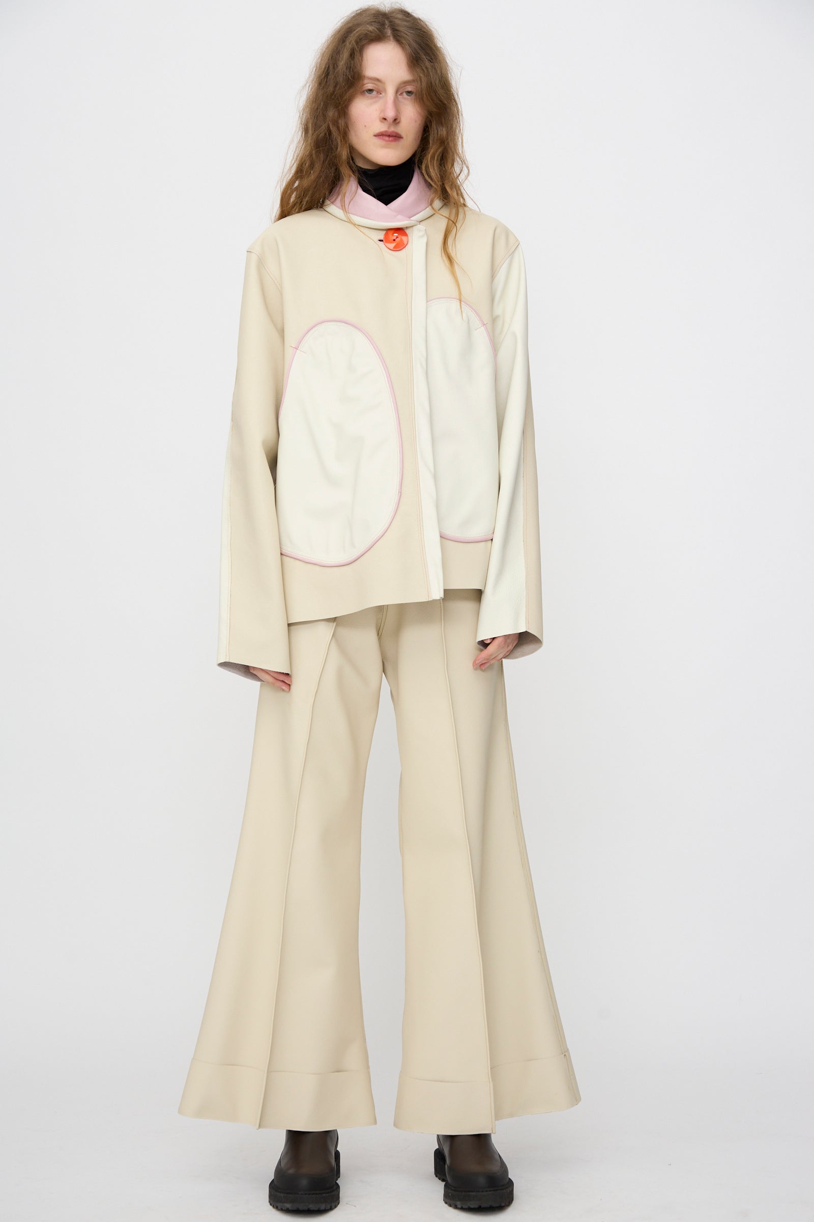 A person stands against a plain background wearing the Leather Chore Jacket in Ecru by Thank You Have A Good Day—handmade in NYC—paired with wide-leg pants and black shoes.