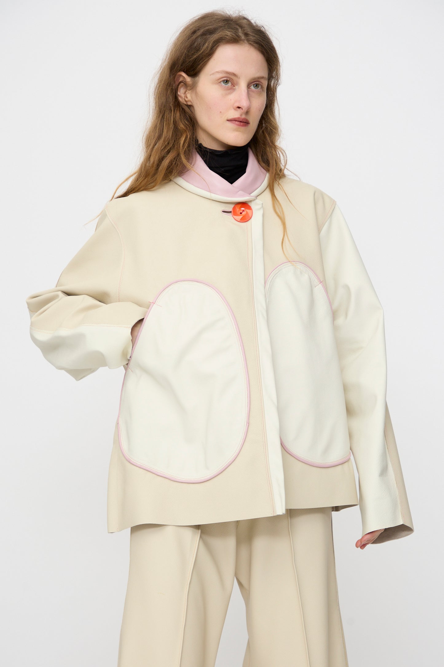 Person wearing the Thank You Have A Good Day Leather Chore Jacket in Ecru with large white pockets, paired with cream trousers, standing against a plain light background.