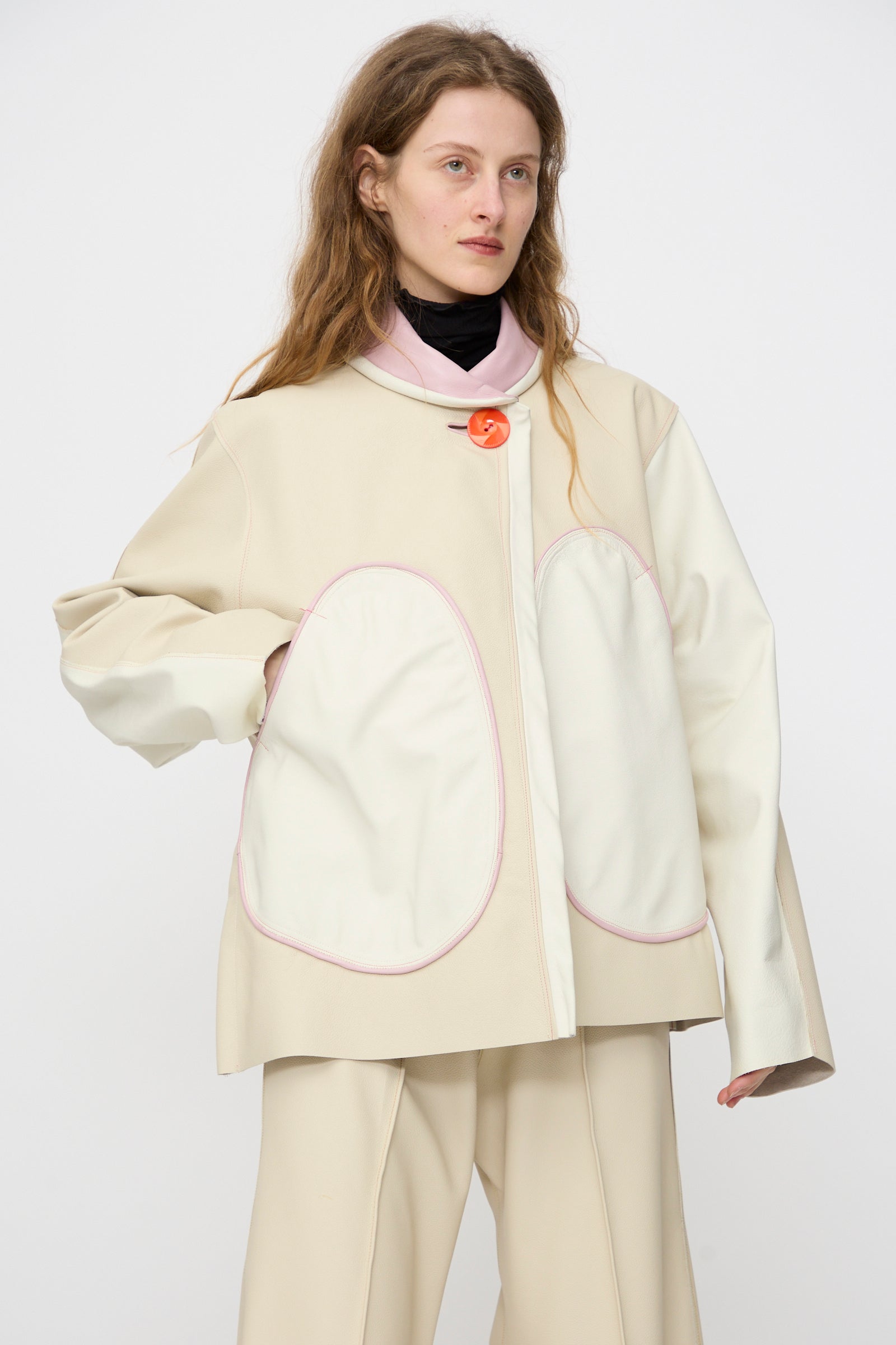 Person wearing the Thank You Have A Good Day Leather Chore Jacket in Ecru with large white pockets, paired with cream trousers, standing against a plain light background.