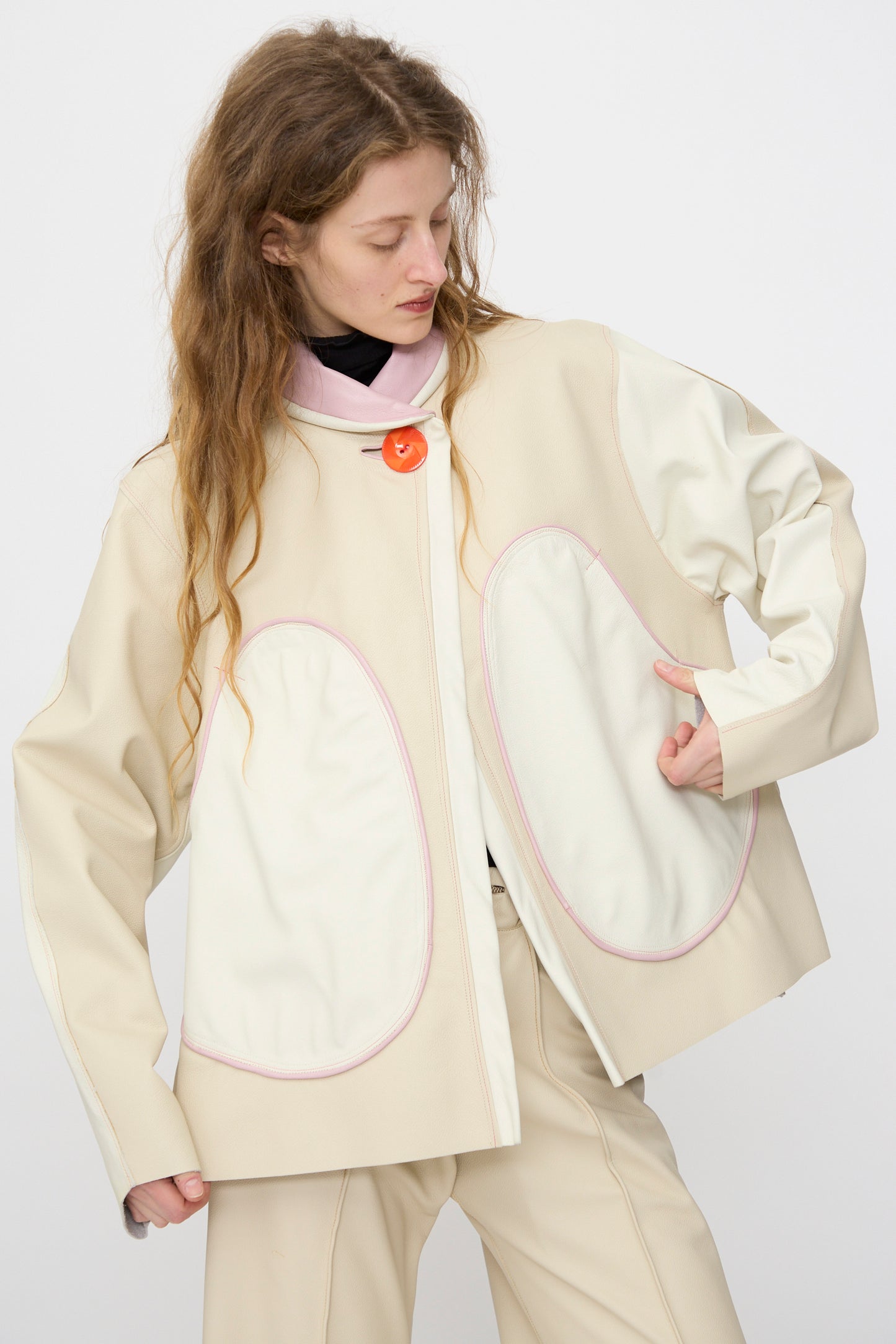 A person wears Thank You Have A Good Day's Leather Chore Jacket in Ecru, featuring large white oval pockets with pink outlines and a single orange button, paired with matching pants.