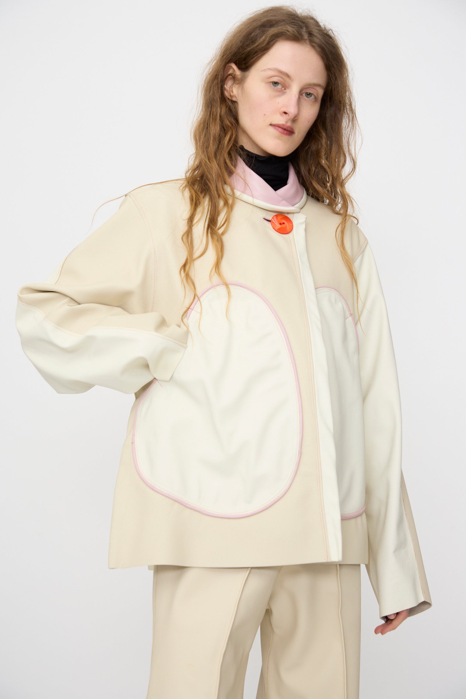 A person with long hair wears the Leather Chore Jacket in Ecru by Thank You Have A Good Day, with oversized white pockets, a pink collar, and a single orange button—handmade in NYC.