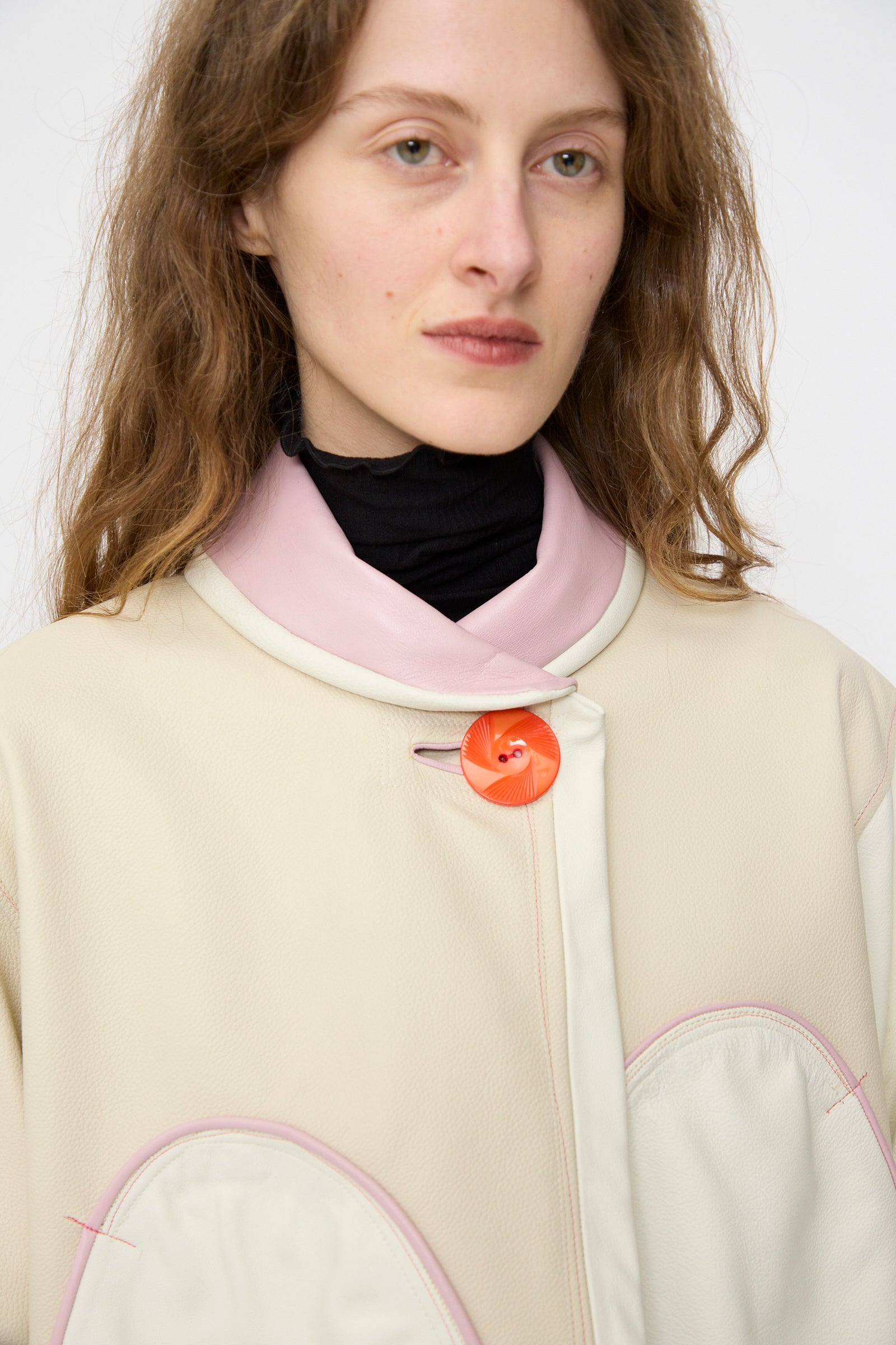 A person with wavy brown hair wears the Thank You Have A Good Day Leather Chore Jacket in Ecru with pink trim and a large orange button over a black turtleneck, posed against a plain background.