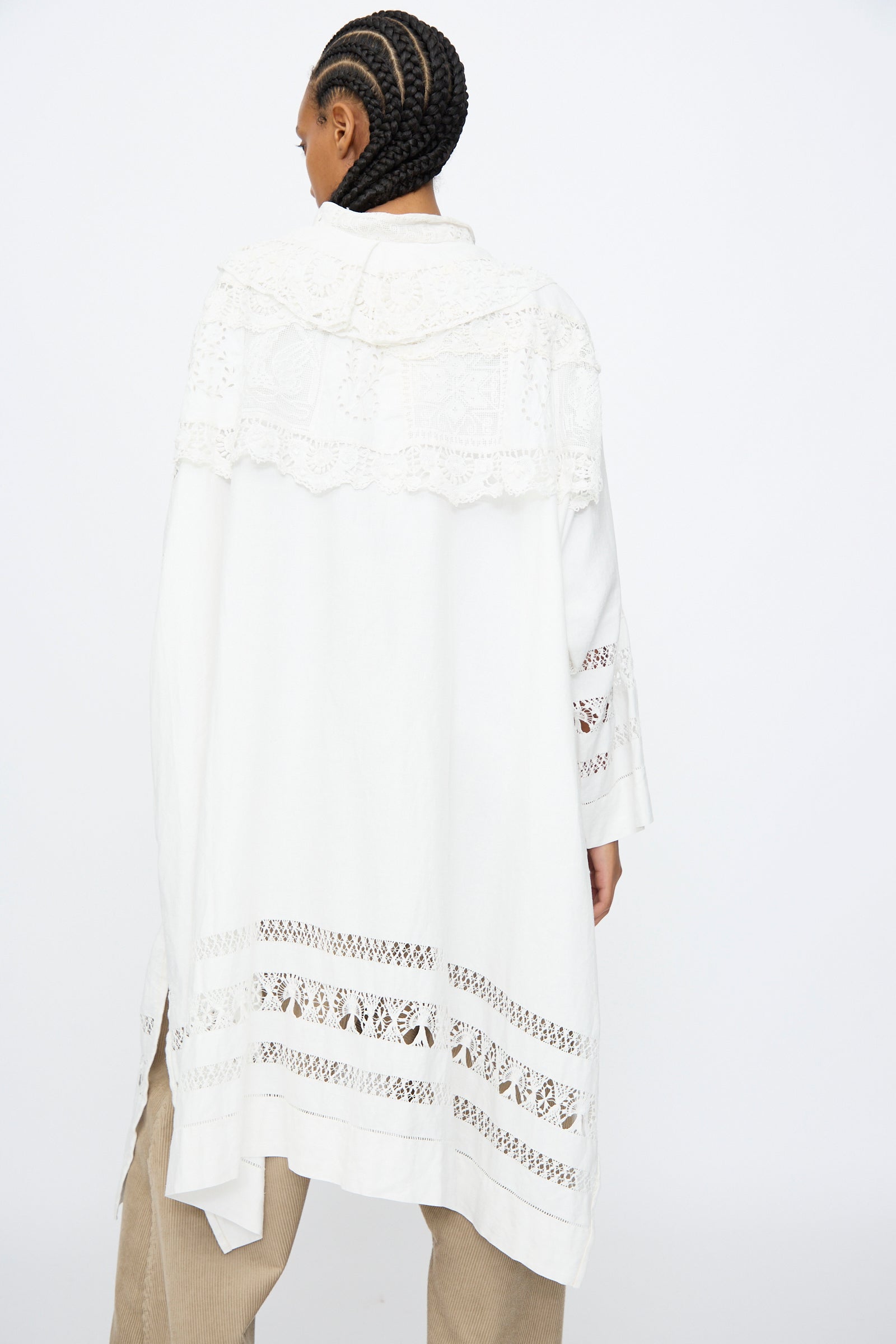Person with braided hair wears the Thank You Have A Good Day Ruffle Oversized Oxford, a long white garment with lace detailing over beige pants, shown from the back against a plain white background.