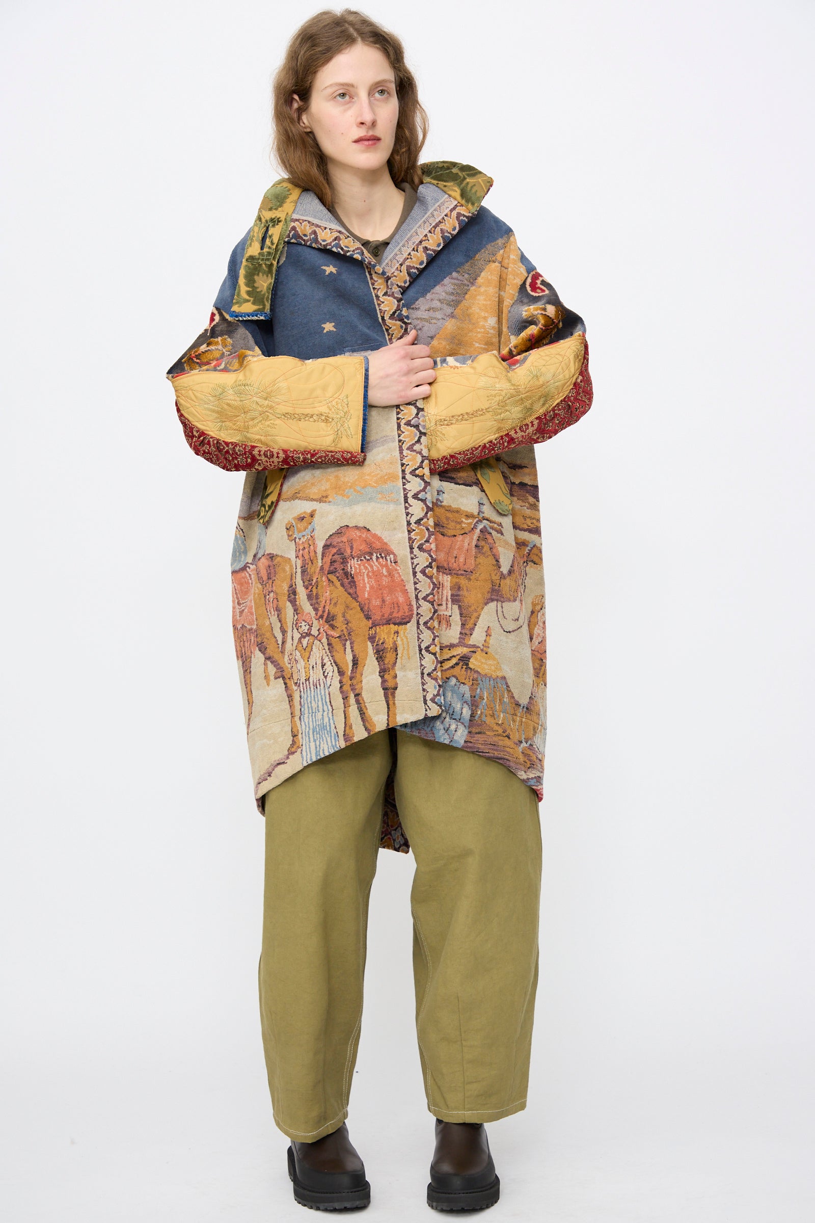A person stands against a plain background in the oversized, colorful Scenic Tapestry Fishtail Parka by Thank You Have A Good Day, featuring animal patterns crafted from repurposed tapestry, paired with olive green pants and black shoes.