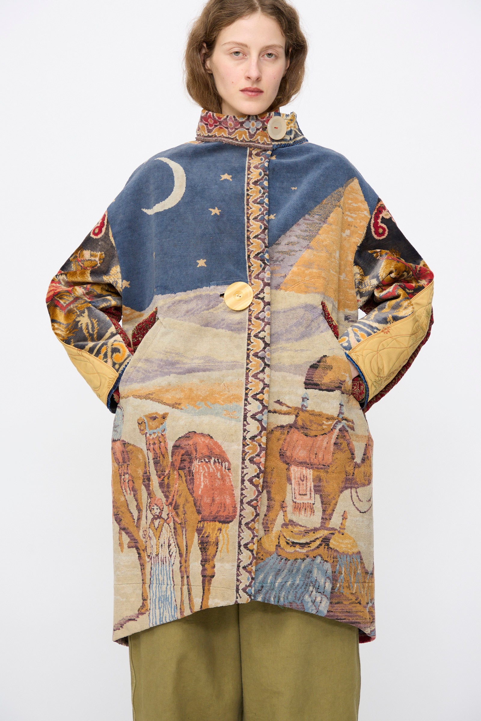 A person models the Scenic Tapestry Fishtail Parka by Thank You Have A Good Day— a handmade, loose parka with desert, camel, and night sky patterns, crafted from repurposed tapestry and featuring large buttons and wide sleeves.