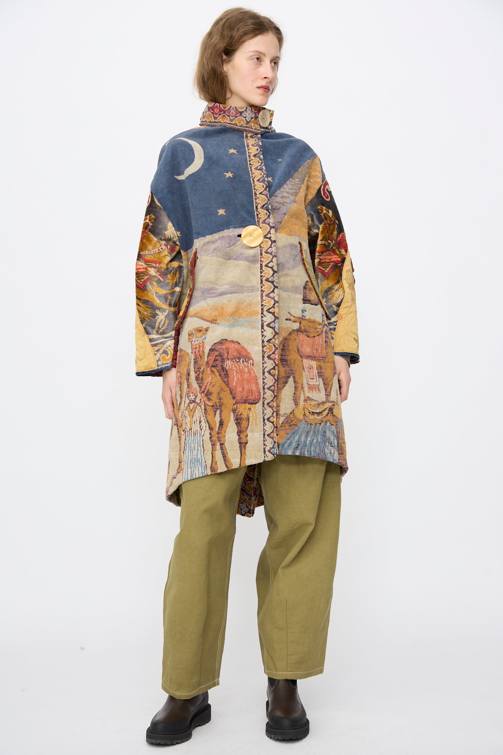A person wearing the Thank You Have A Good Day Scenic Tapestry Fishtail Parka, featuring desert and night sky designs, along with olive green pants and dark boots, stands against a plain white background.