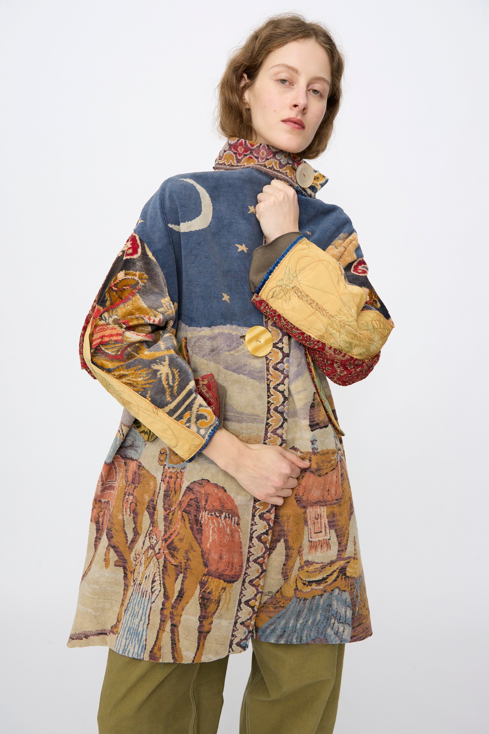 A person wears the Scenic Tapestry Fishtail Parka by Thank You Have A Good Day, featuring moons, stars, and camels, standing against a plain light background.