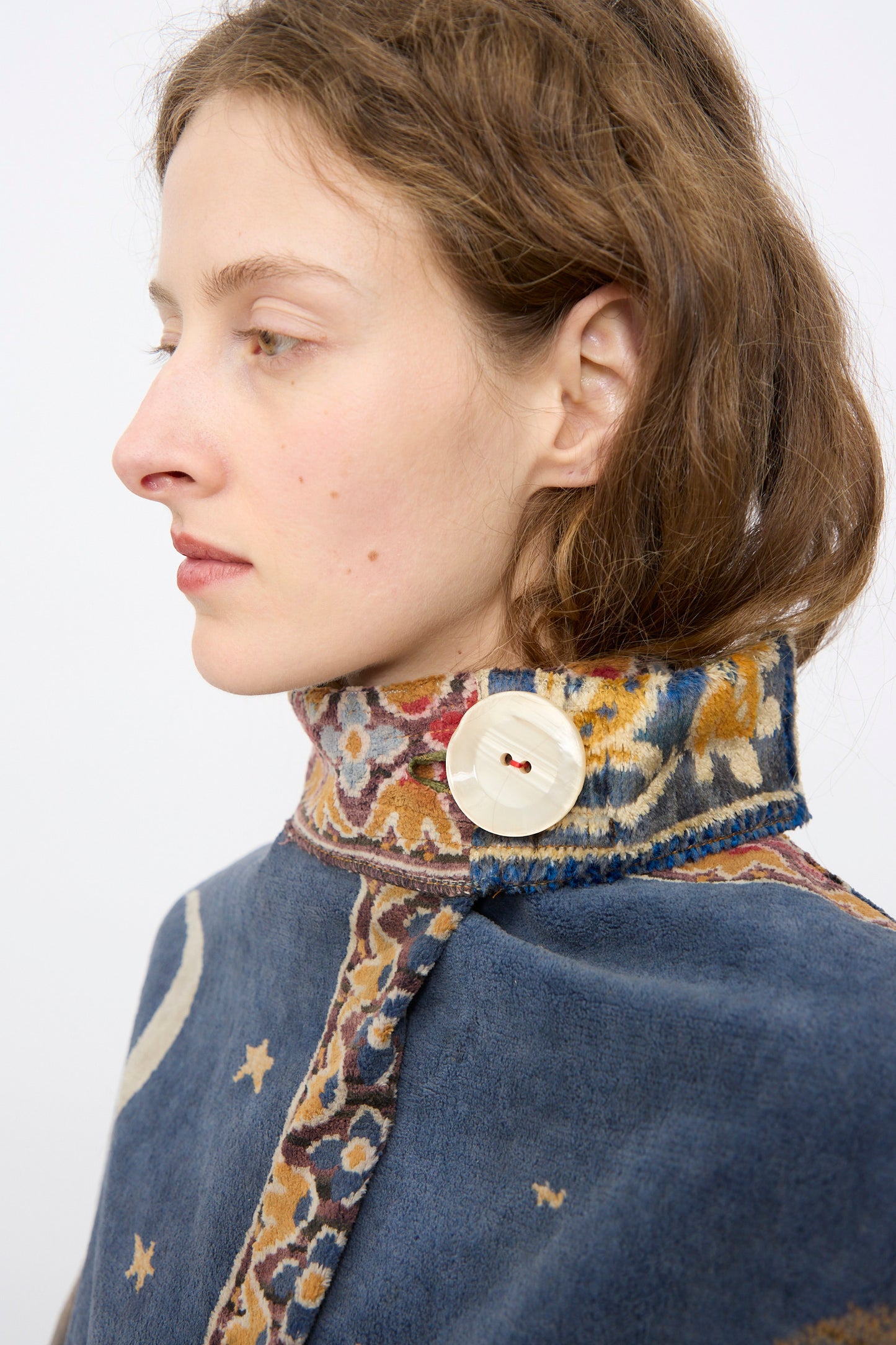 A woman in profile wears the Scenic Tapestry Fishtail Parka by Thank You Have A Good Day, featuring a high collar, large decorative button, and patterned trim with stars and a crescent moon.