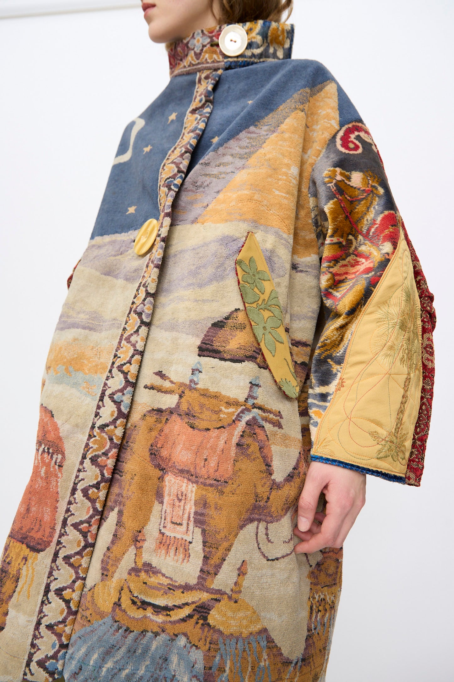 A person wears the Scenic Tapestry Fishtail Parka by Thank You Have A Good Day, featuring tapestry-like ancient Egyptian motifs, decorative buttons, and crafted from repurposed tapestry.
