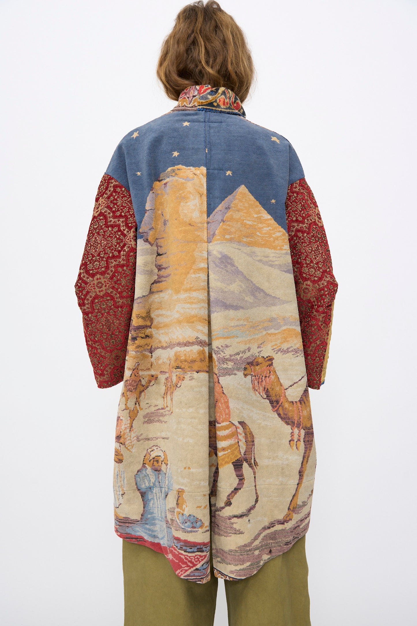 A person with short brown hair stands with their back to the camera, wearing the "Scenic Tapestry Fishtail Parka" by Thank You Have A Good Day, featuring a desert scene with pyramids, camels, figures, and patterned red sleeves.