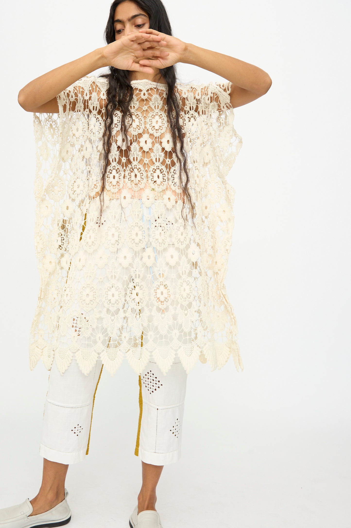 A person wears the Antique Lace Midi Caftan by Thank You Have A Good Day, its cream hue and crochet lace details layered over white cropped pants with cut-out patterns, standing against a plain backdrop with arms partially covering their face.