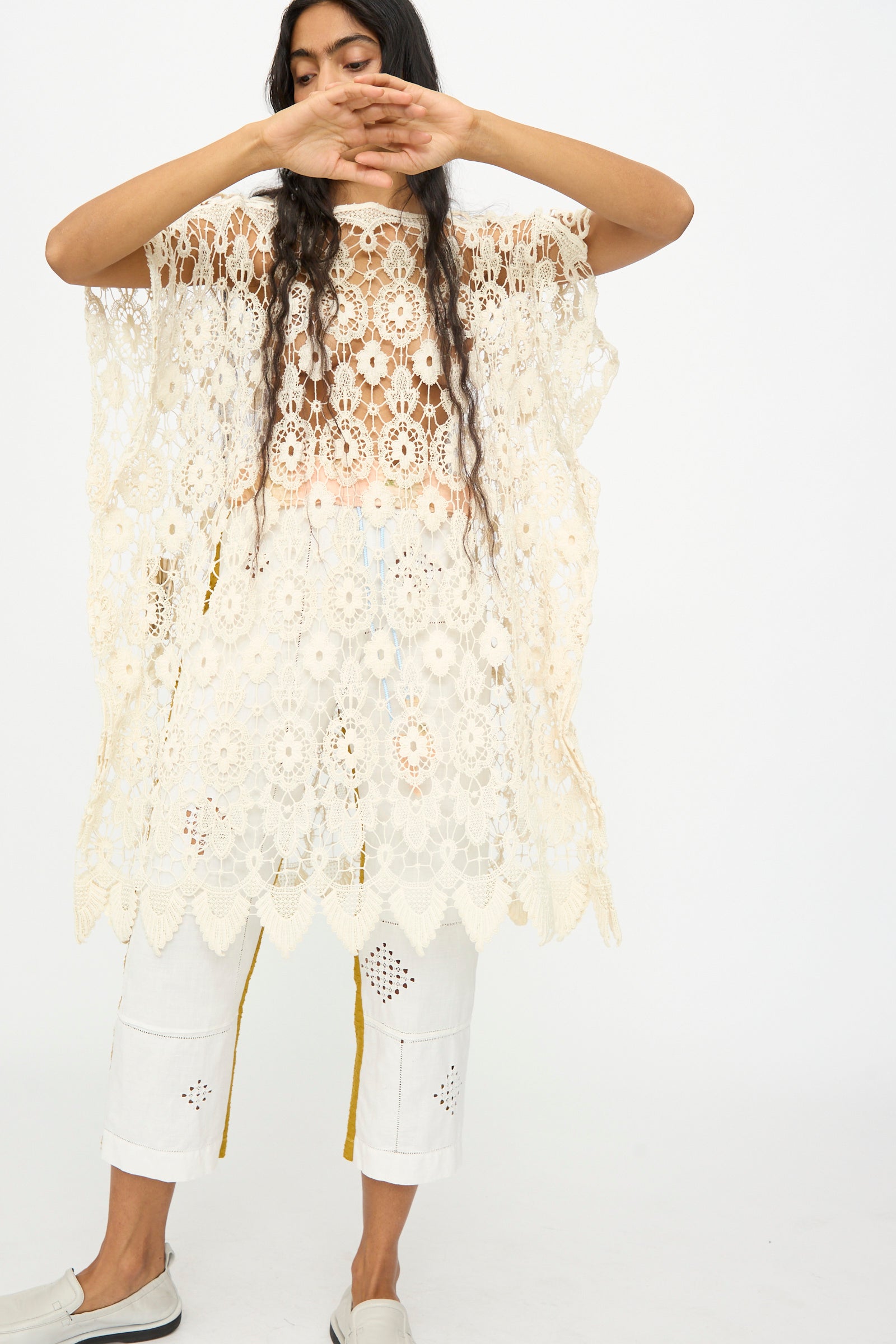 A person wears the Antique Lace Midi Caftan by Thank You Have A Good Day, its cream hue and crochet lace details layered over white cropped pants with cut-out patterns, standing against a plain backdrop with arms partially covering their face.