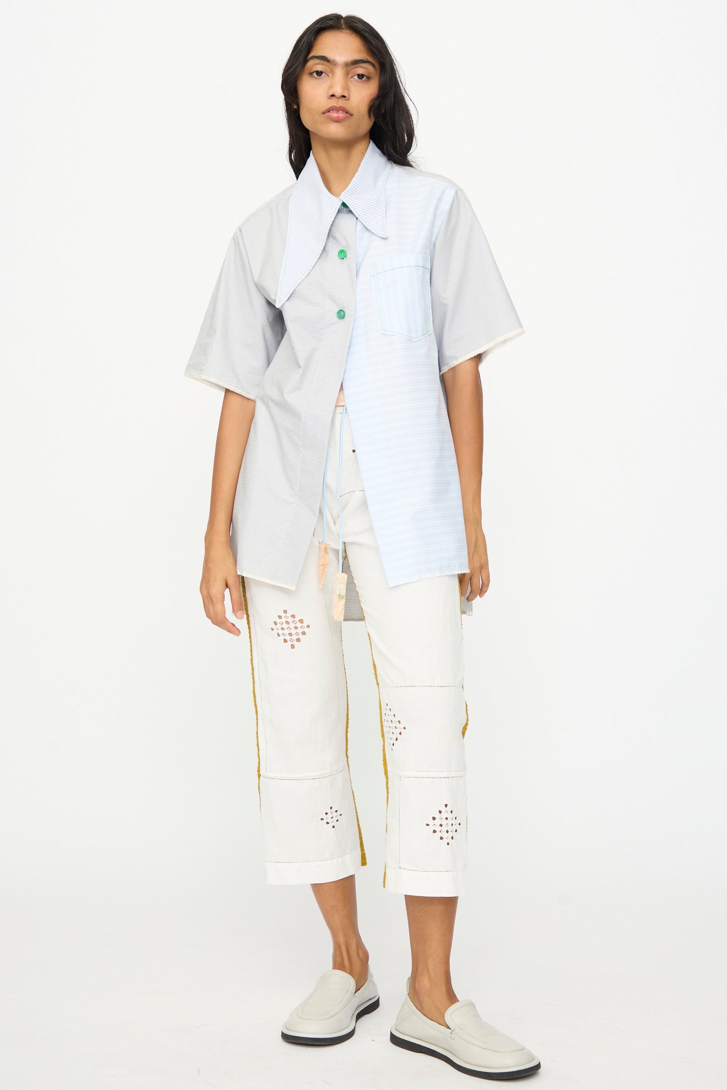 Against a plain background, a person wears Thank You Have A Good Day’s Asymmetrical Collar Shirt with mixed blue and gray panels, cropped white cut-out pants handmade in Tribeca, and white slip-on shoes.