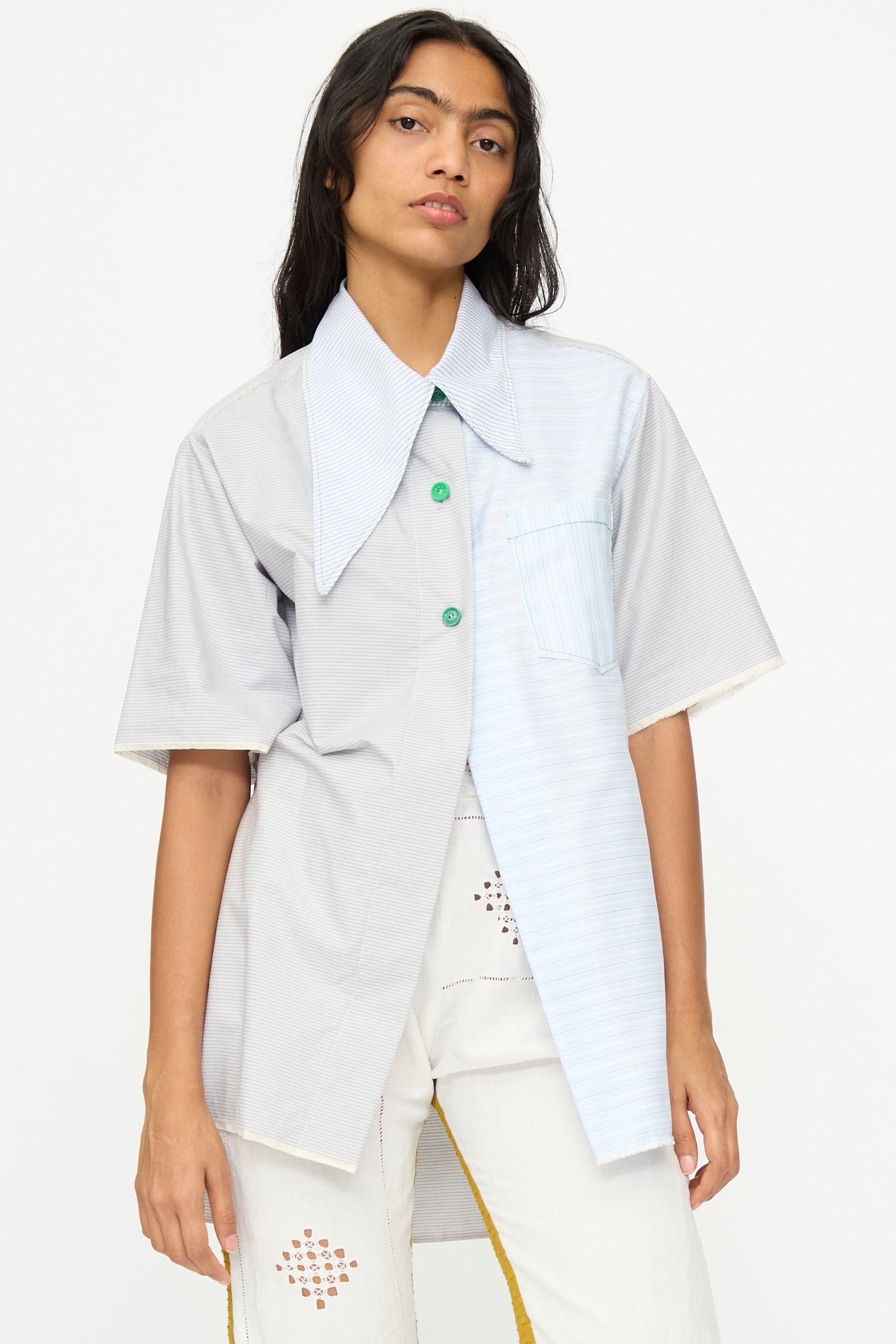 A woman wears the Asymmetrical Collar Shirt by Thank You Have A Good Day—featuring a two-tone design, oversized collar, green buttons, and striped cotton—paired with white pants with cutout details.