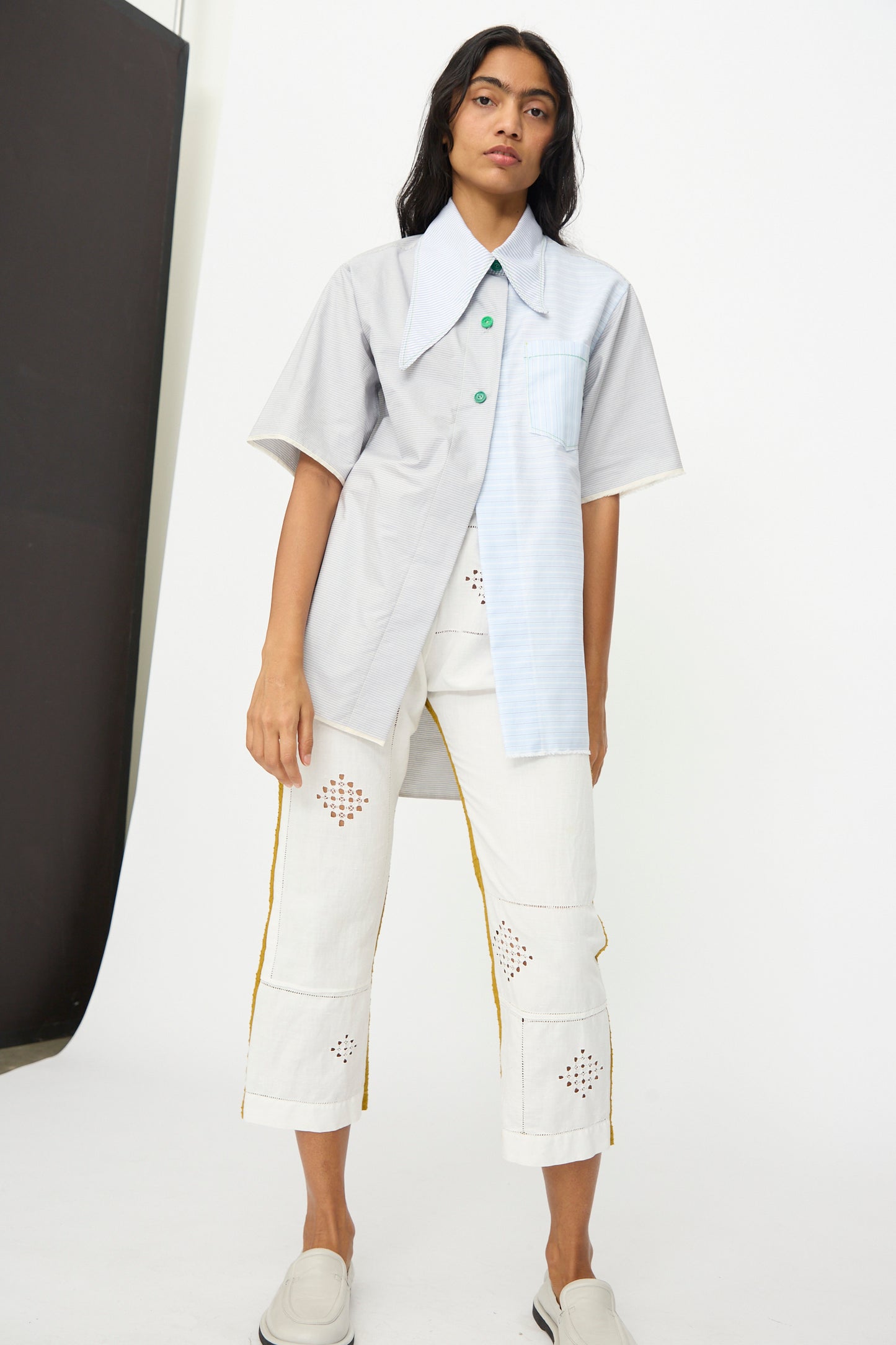 A person wears the Asymmetrical Collar Shirt by Thank You Have A Good Day, a light gray, handmade short-sleeve oxford with mismatched panels, paired with white cropped pants featuring geometric cutouts and yellow side stripes.
