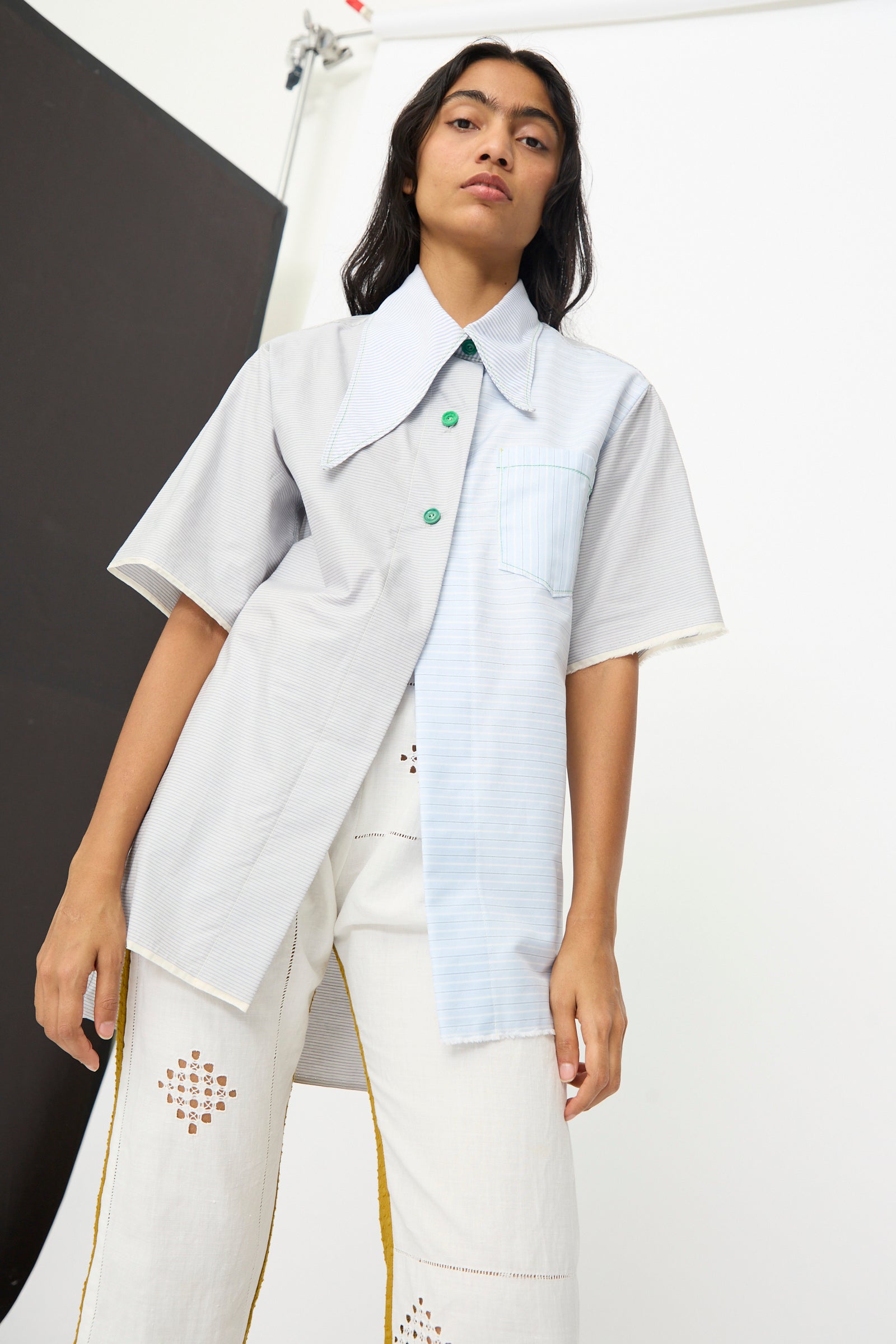 A person wears the Thank You Have A Good Day Asymmetrical Collar Shirt—short-sleeve, two-tone, with green buttons—and white cutout pants, standing against a minimal studio background.