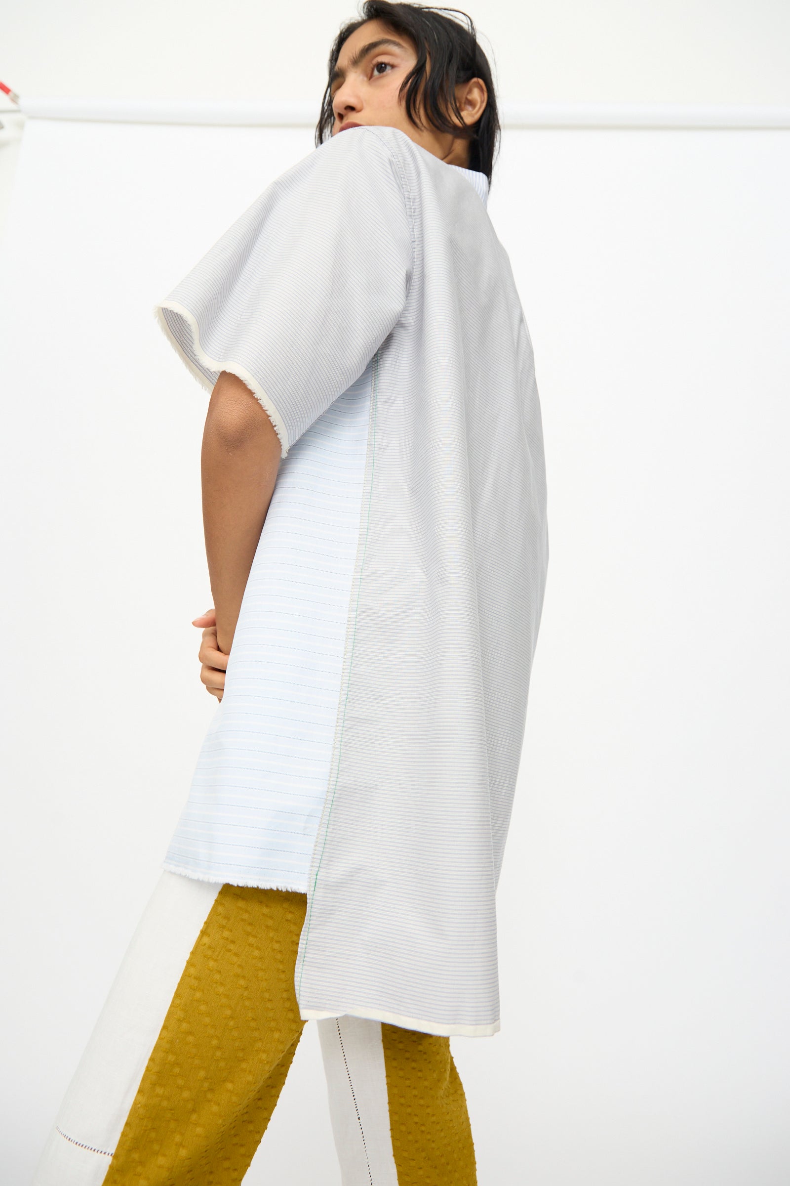 A person wears the Thank You Have A Good Day Asymmetrical Collar Shirt, a loose, short-sleeved light gray top, paired with striped yellow and white cotton pants, standing before a plain white background.