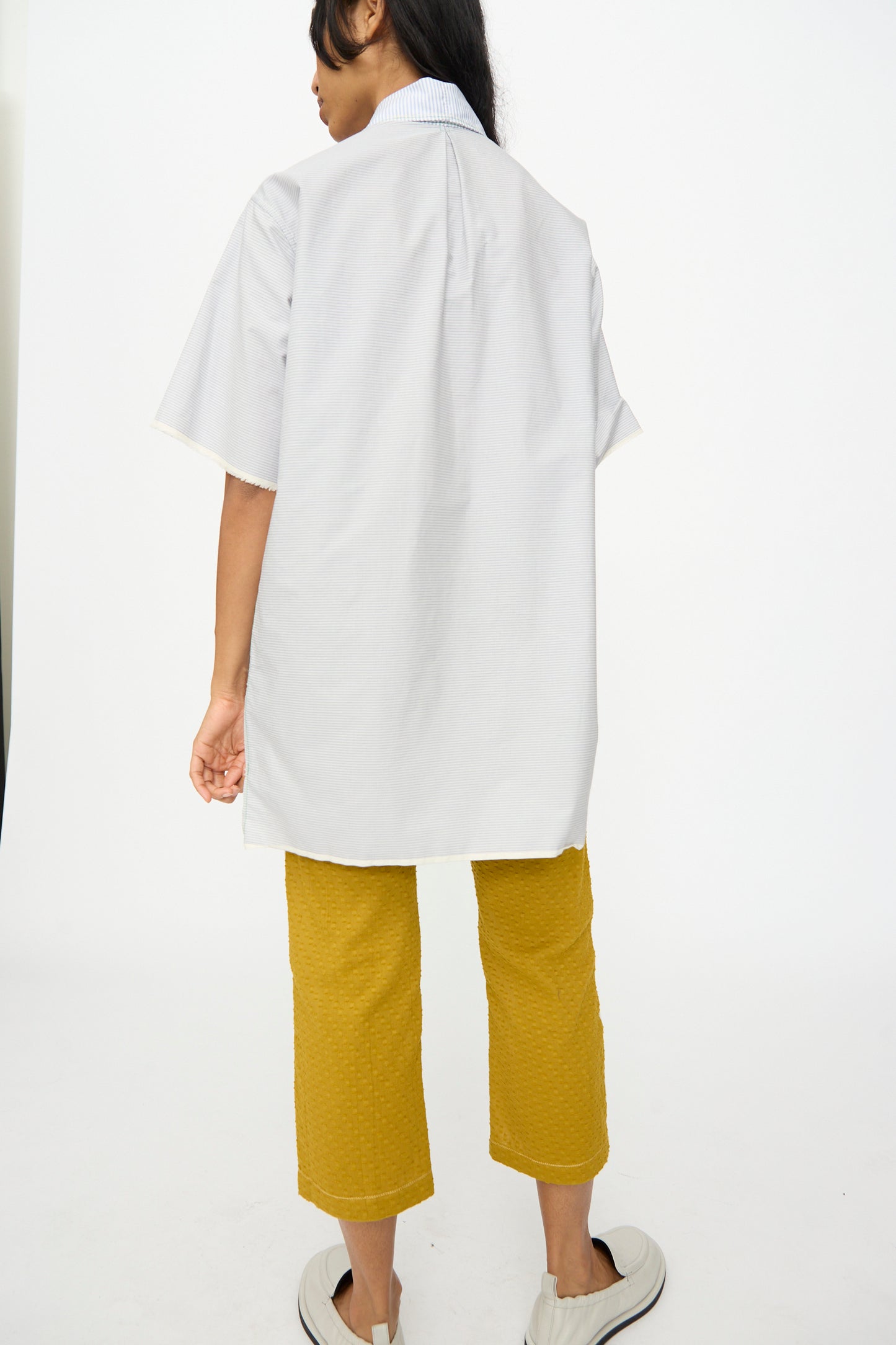Person stands against a white background, facing away, wearing Thank You Have A Good Day's Asymmetrical Collar Shirt in light gray short sleeves, yellow cropped pants, and white slip-on shoes.