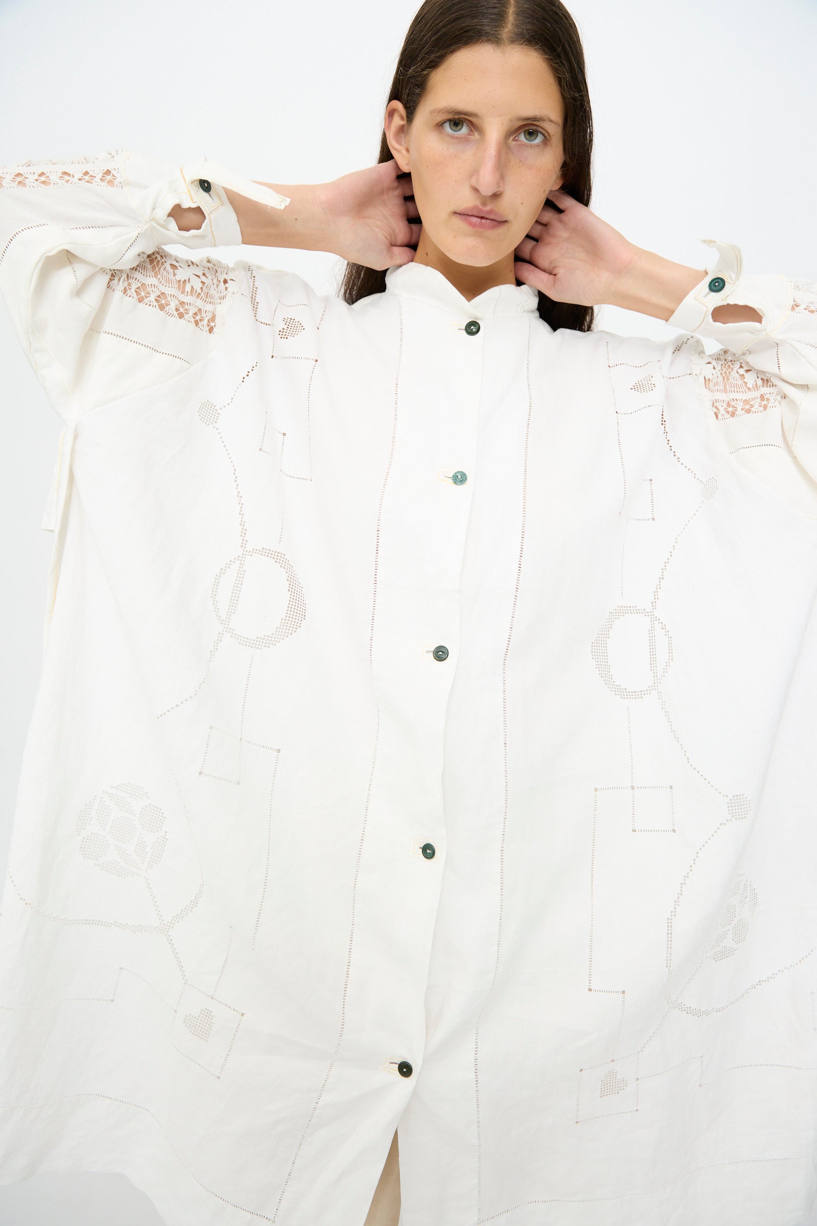 A woman with long brown hair models the Thank You Have A Good Day Heart and Flower Oversized Oxford shirt dress, featuring geometric patterns and lace details, posing with hands behind her neck against a plain white background.