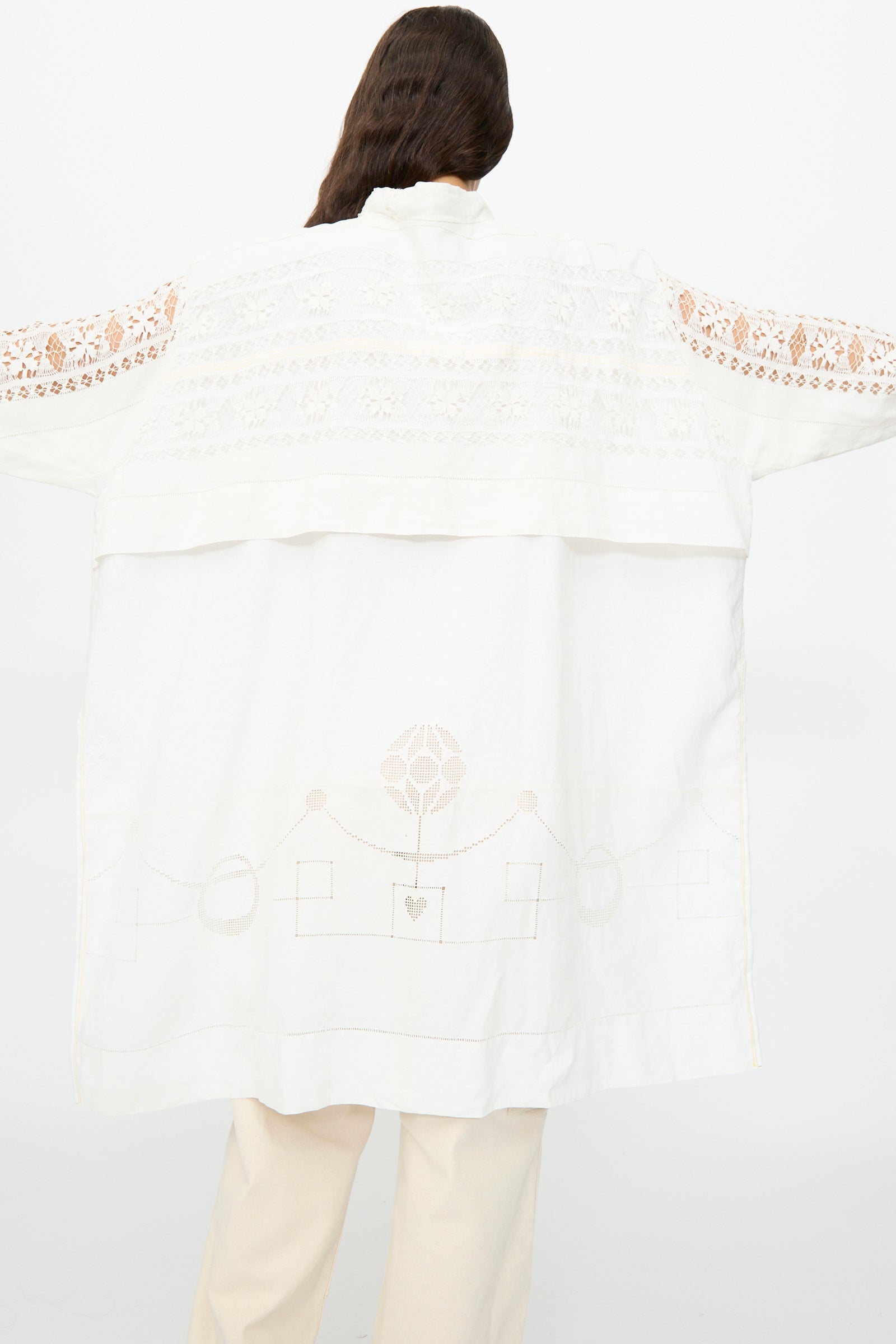 A person with long brown hair models the Thank You Have A Good Day Heart and Flower Oversized Oxford shirt dress in white, featuring delicate lace details, arms outstretched, viewed from the back.