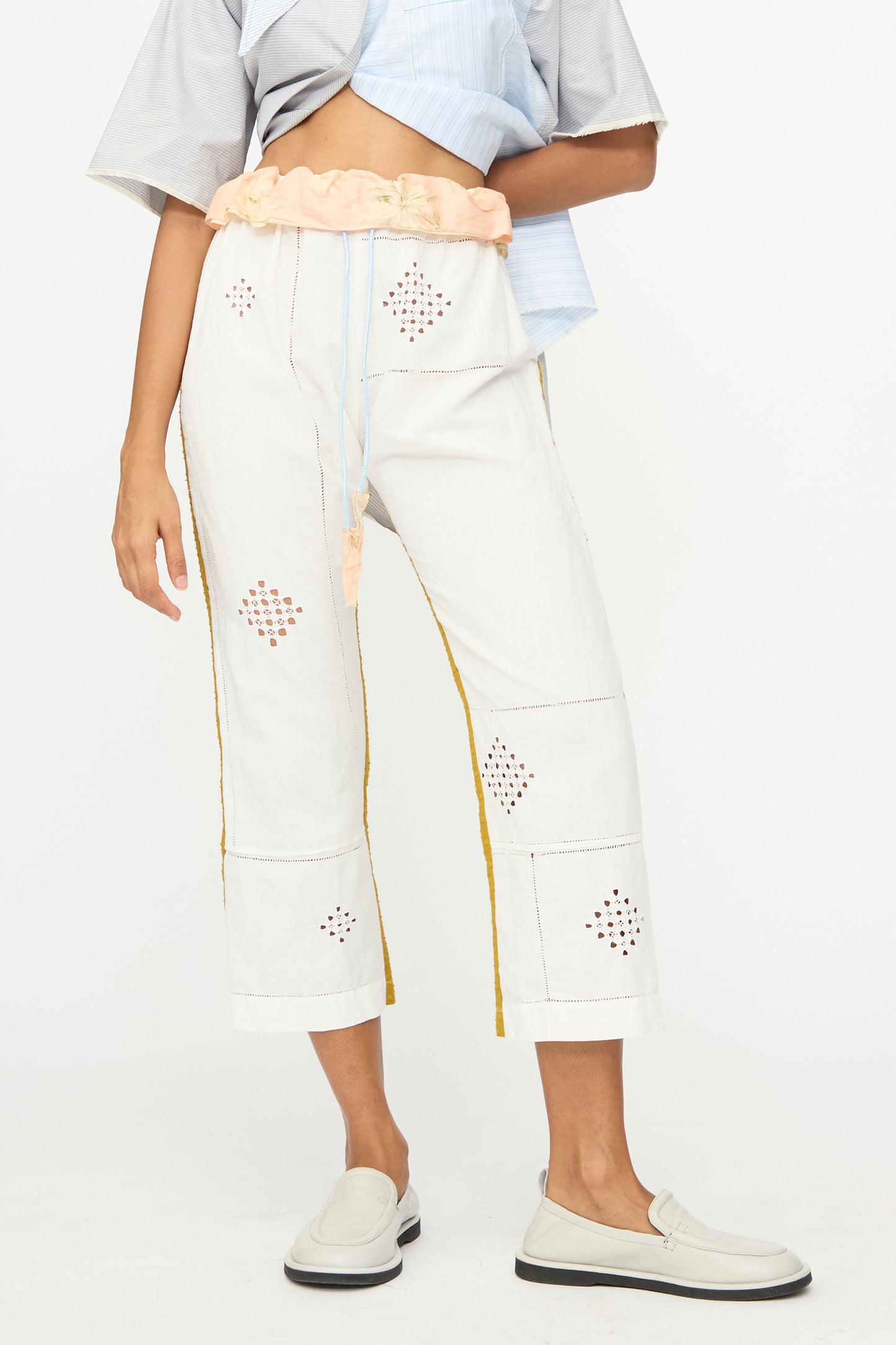 A person wears the Lounge Pant by Thank You Have A Good Day—cropped white pants with decorative cutouts and a peach elastic waistband—paired with a gray top and white loafers, standing against a plain background.