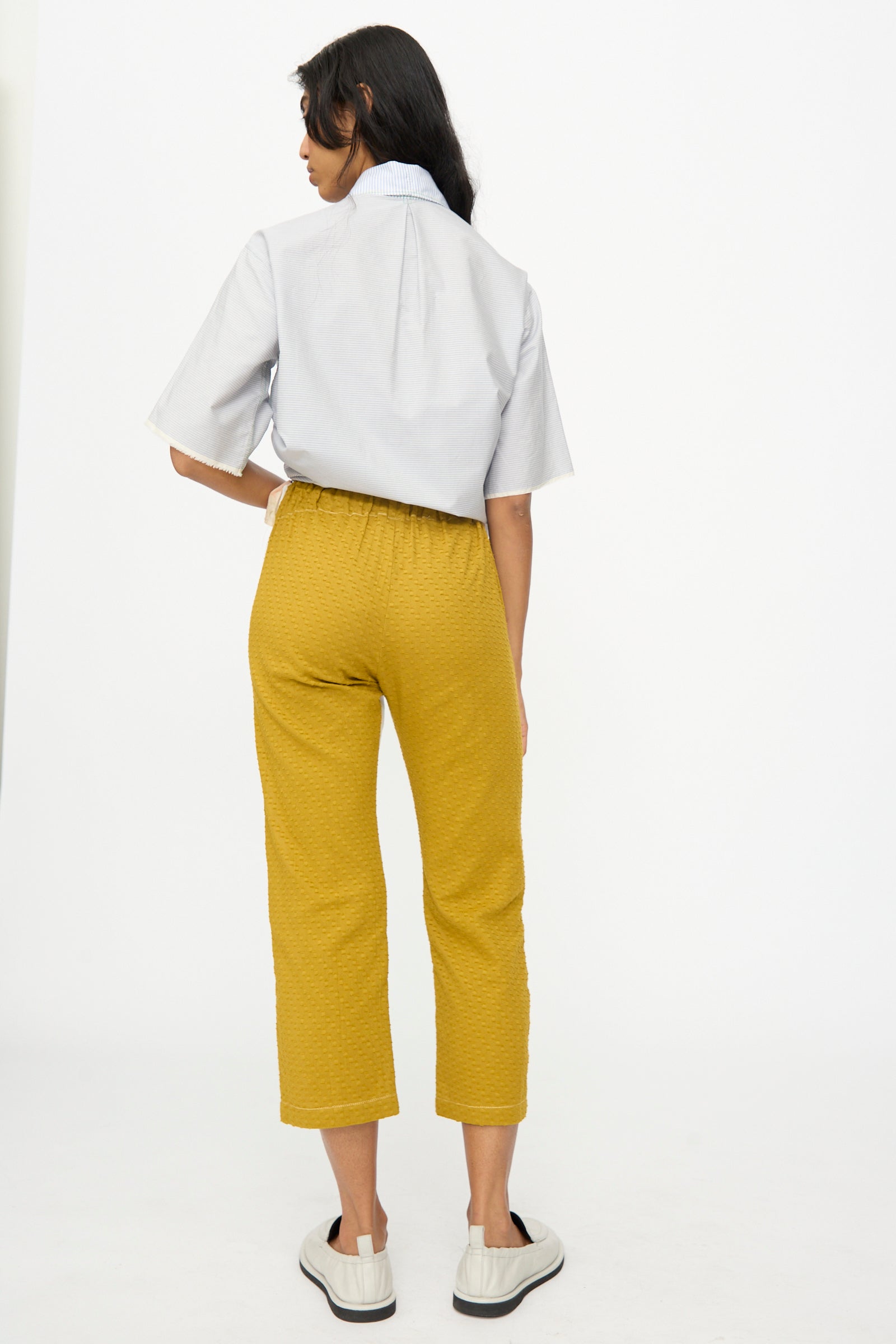 A woman stands with her back to the camera, wearing a light blue short-sleeve shirt and Thank You Have A Good Day Lounge Pant in mustard yellow, paired with white slip-on shoes against a plain white background.