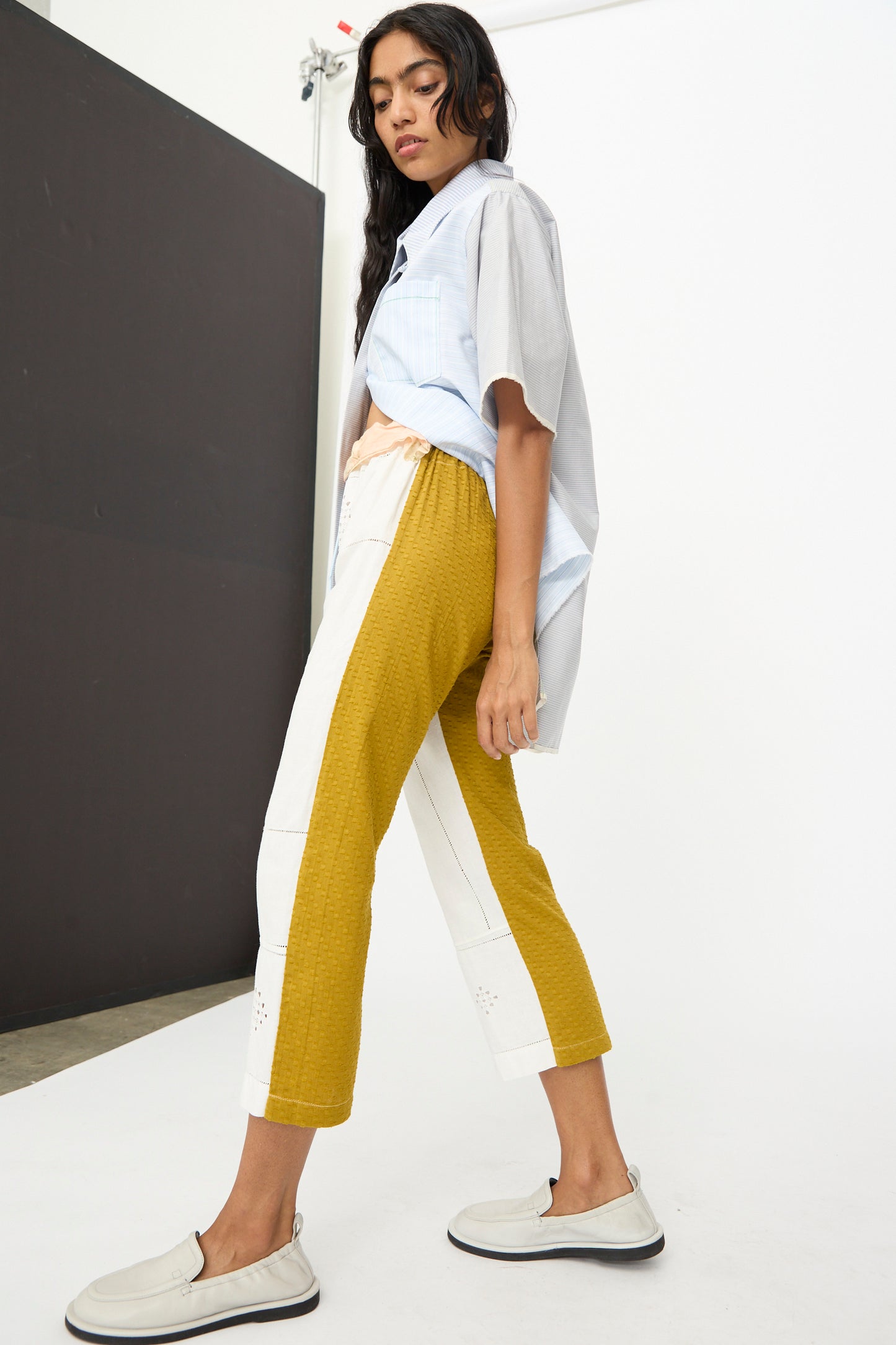 A woman stands in profile wearing the Thank You Have A Good Day Lounge Pant in colorblock yellow and white, paired with a loose short-sleeve shirt and slip-on shoes against a minimal studio background.