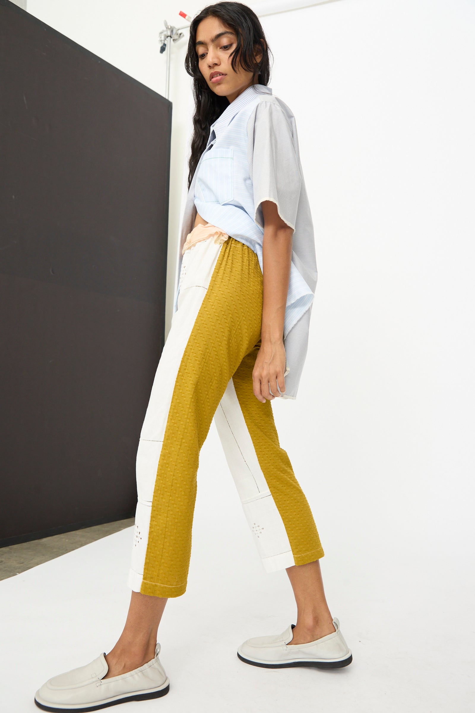 A woman stands in profile wearing the Thank You Have A Good Day Lounge Pant in colorblock yellow and white, paired with a loose short-sleeve shirt and slip-on shoes against a minimal studio background.