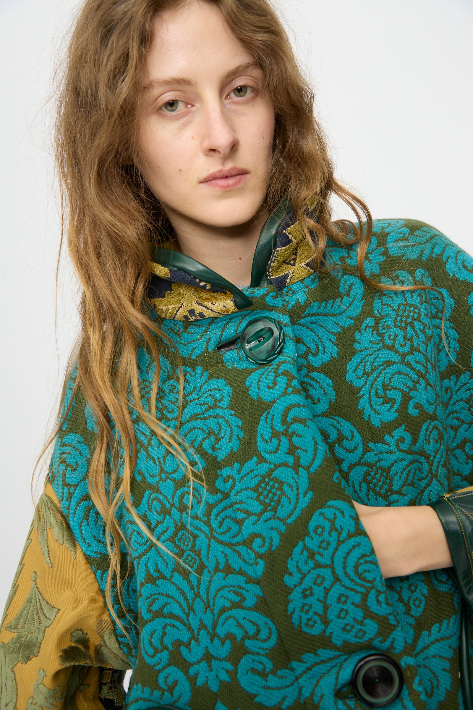 A person with long wavy hair wears the Patchwork Fishtail Parka by Thank You Have A Good Day, featuring green and blue patterns, vintage button closure, gold sleeve details, and antique tapestry-inspired design against a white background.