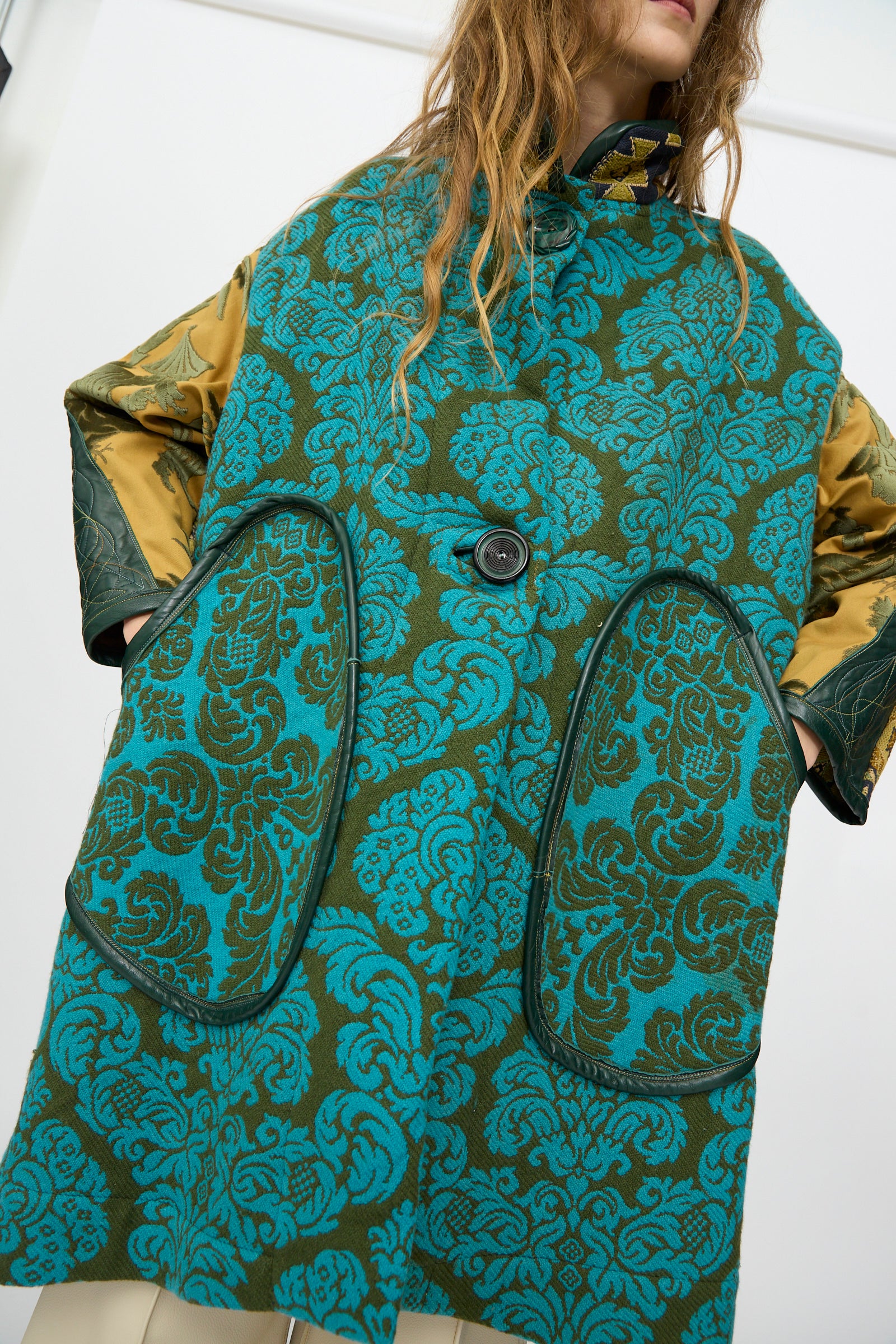 A person wears the Patchwork Fishtail Parka by Thank You Have A Good Day, featuring a green and blue ornate pattern, large pockets, gold sleeves, vintage button closure, and crafted from antique tapestry textiles against a white background.
