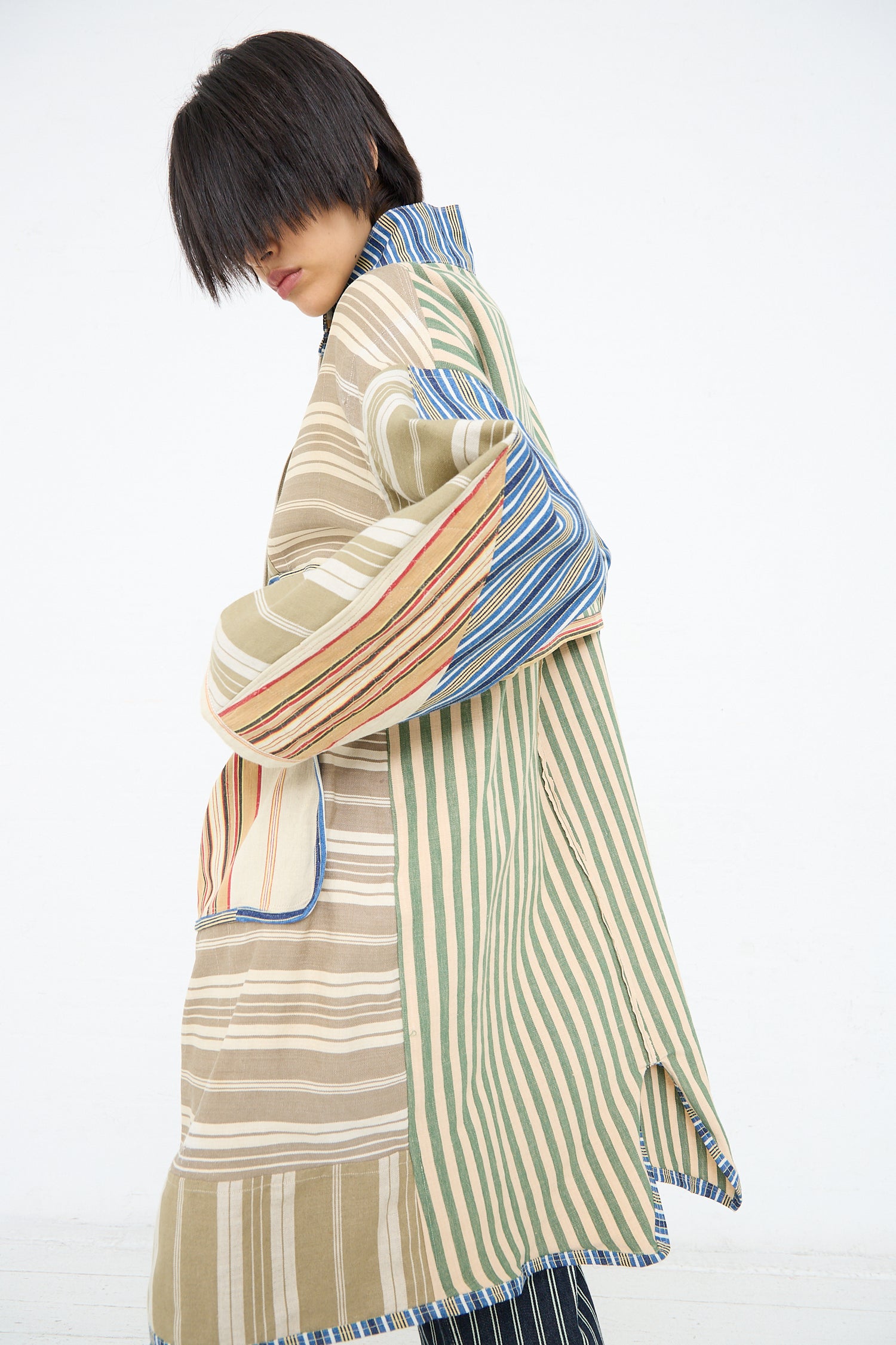 A person is wearing a Thank You Have A Good Day Patchwork French Ticking Fishtail Parka with varying patterns and colors, posing with their back partly towards the camera and looking down to their left.