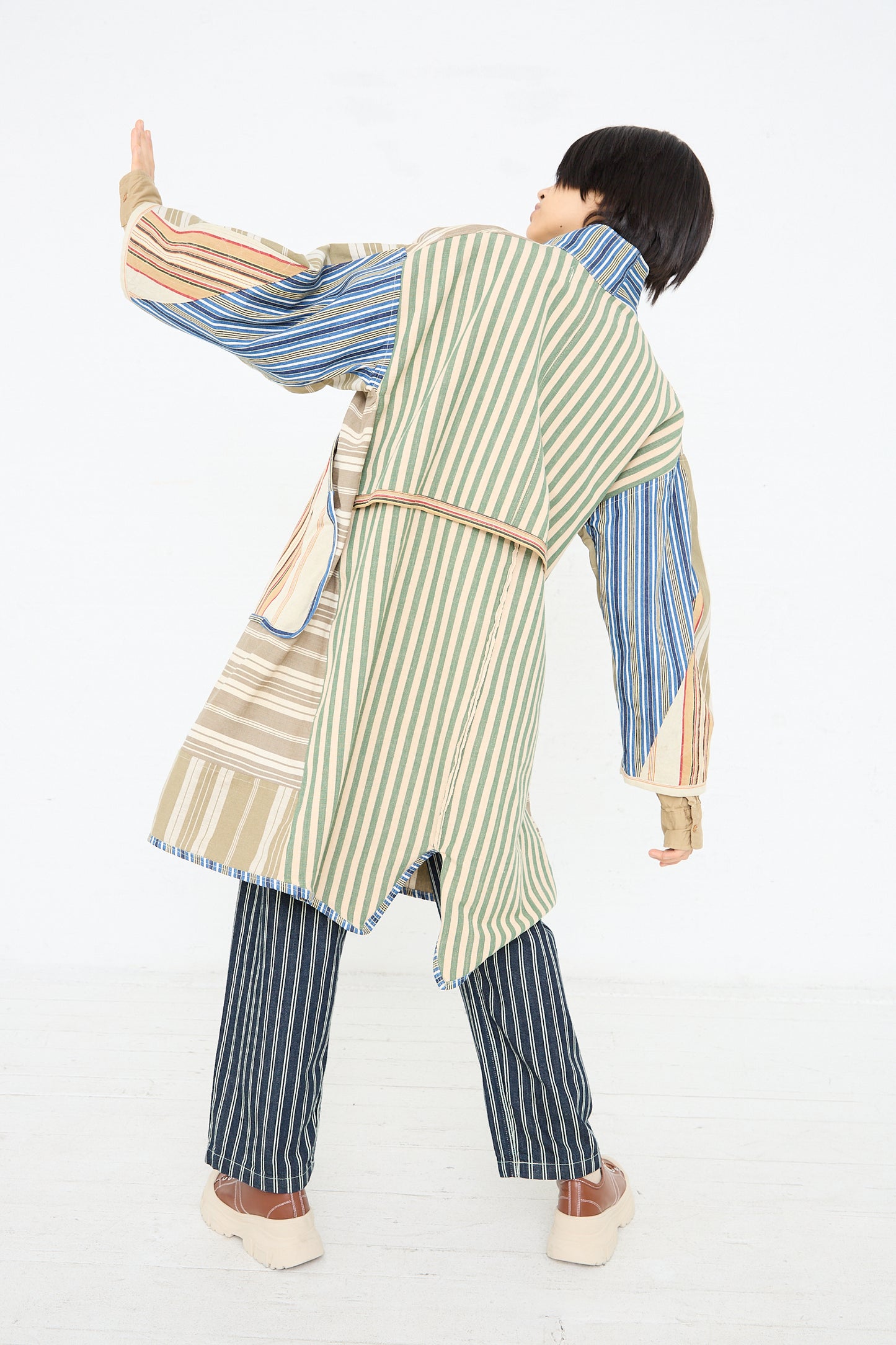 A person wearing a Thank You Have A Good Day Patchwork French Ticking Fishtail Parka with an avant-garde design poses with their back to the camera and one arm extended upwards against a white background.
