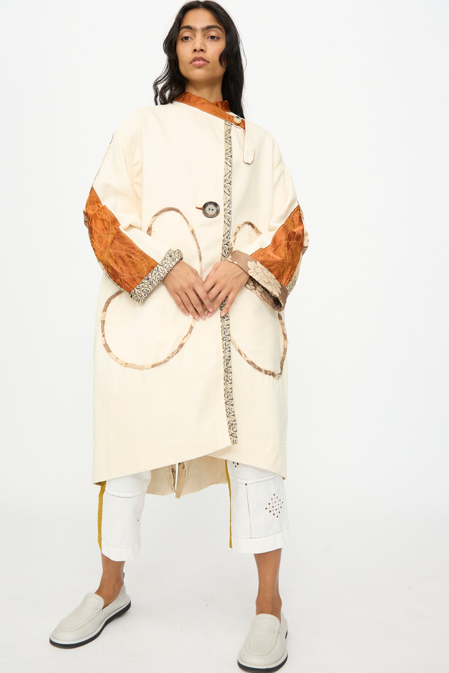 A person models the Thank You Have A Good Day Patchwork Parka, featuring vintage buttons, orange sleeves, and patterned trim, paired with white cropped pants and slip-on shoes against a plain white background.