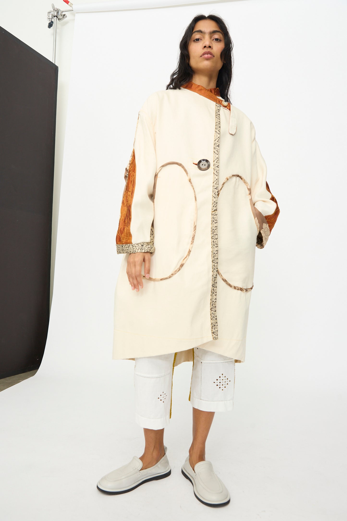 Model wears the Patchwork Parka by Thank You Have A Good Day, a long cream coat with vintage buttons and patterned trim, styled over white cropped pants and slip-on shoes against a plain studio backdrop.