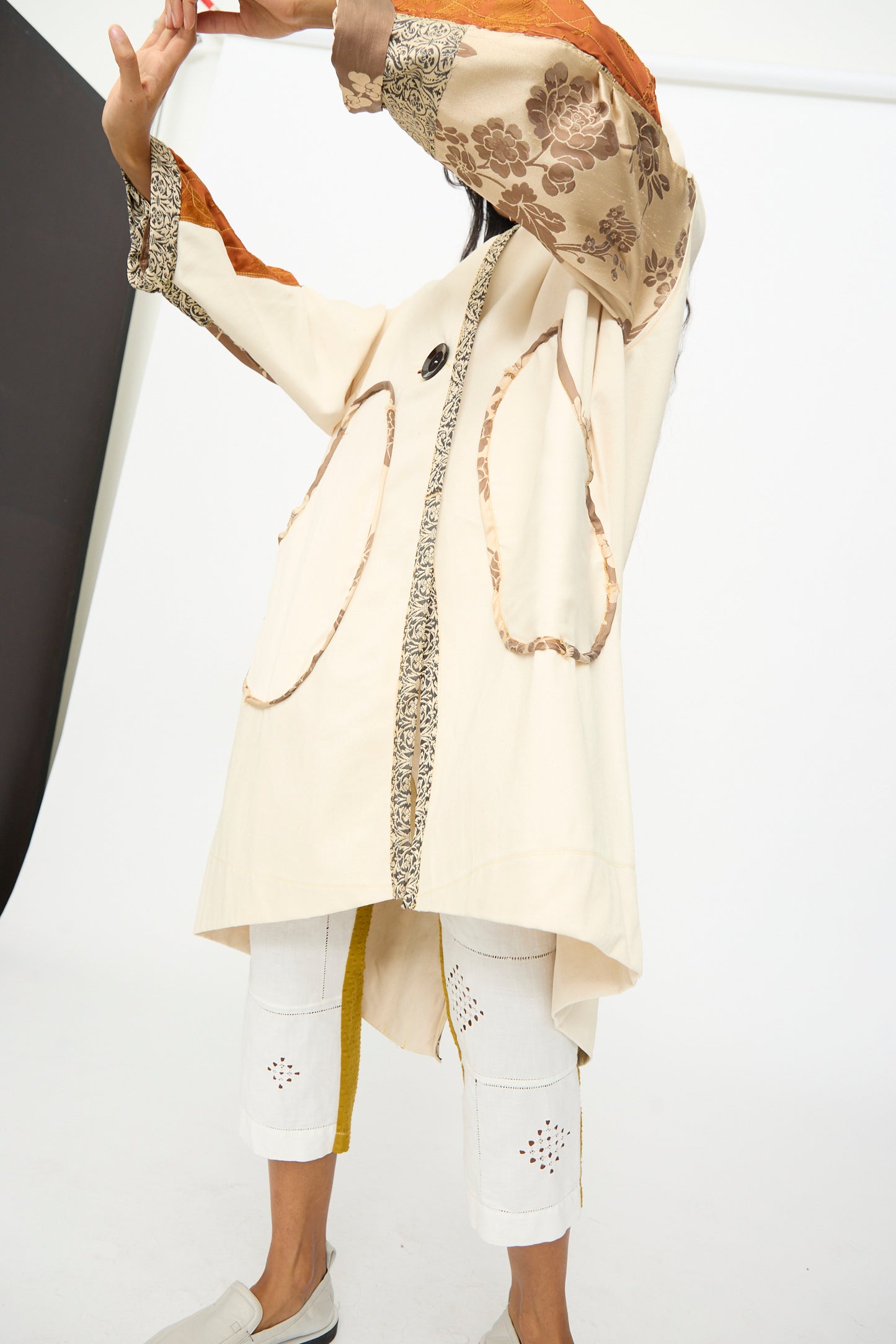 Model wears the Patchwork Parka by Thank You Have A Good Day—featuring patterned sleeves and vintage buttons—paired with white cropped pants with cut-out designs and slip-on shoes, standing against a white backdrop.