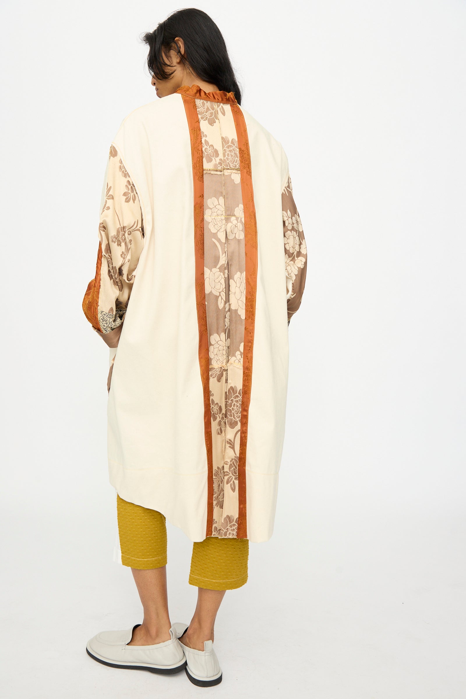 A person faces away in Thank You Have A Good Day’s Patchwork Parka—cream with brown and orange floral panels—paired with yellow cropped pants and light slip-ons, featuring vintage buttons and handmade in Tribeca.