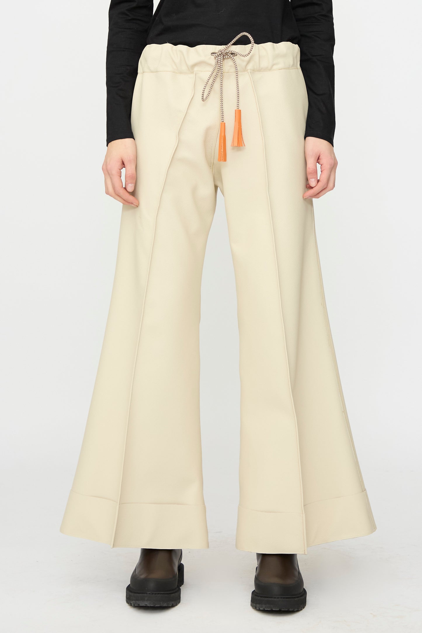 A person wears the Pierrot Flare Trouser in Leather by Thank You Have A Good Day—cream-colored, wide-leg with orange tassels—paired with a black top and boots, standing against a plain background.