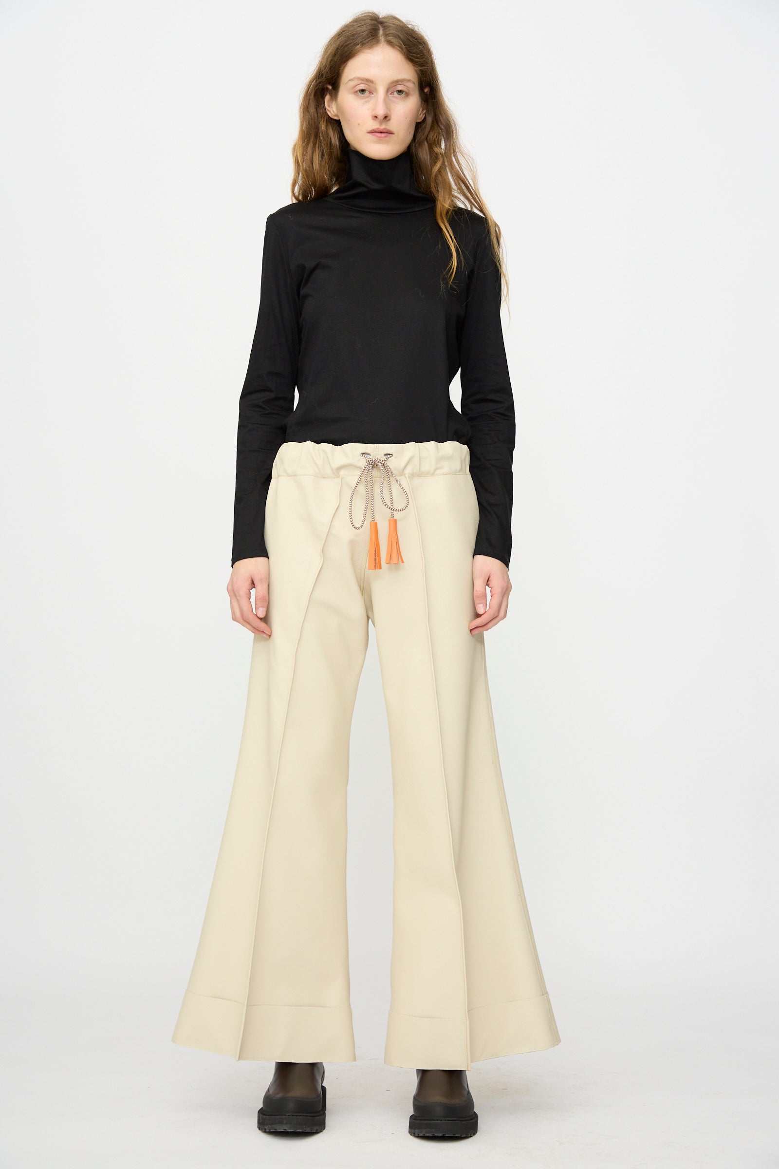A person stands against a plain white background wearing the Thank You Have A Good Day Pierrot Flare Trouser in Leather, styled with a black turtleneck and black shoes.