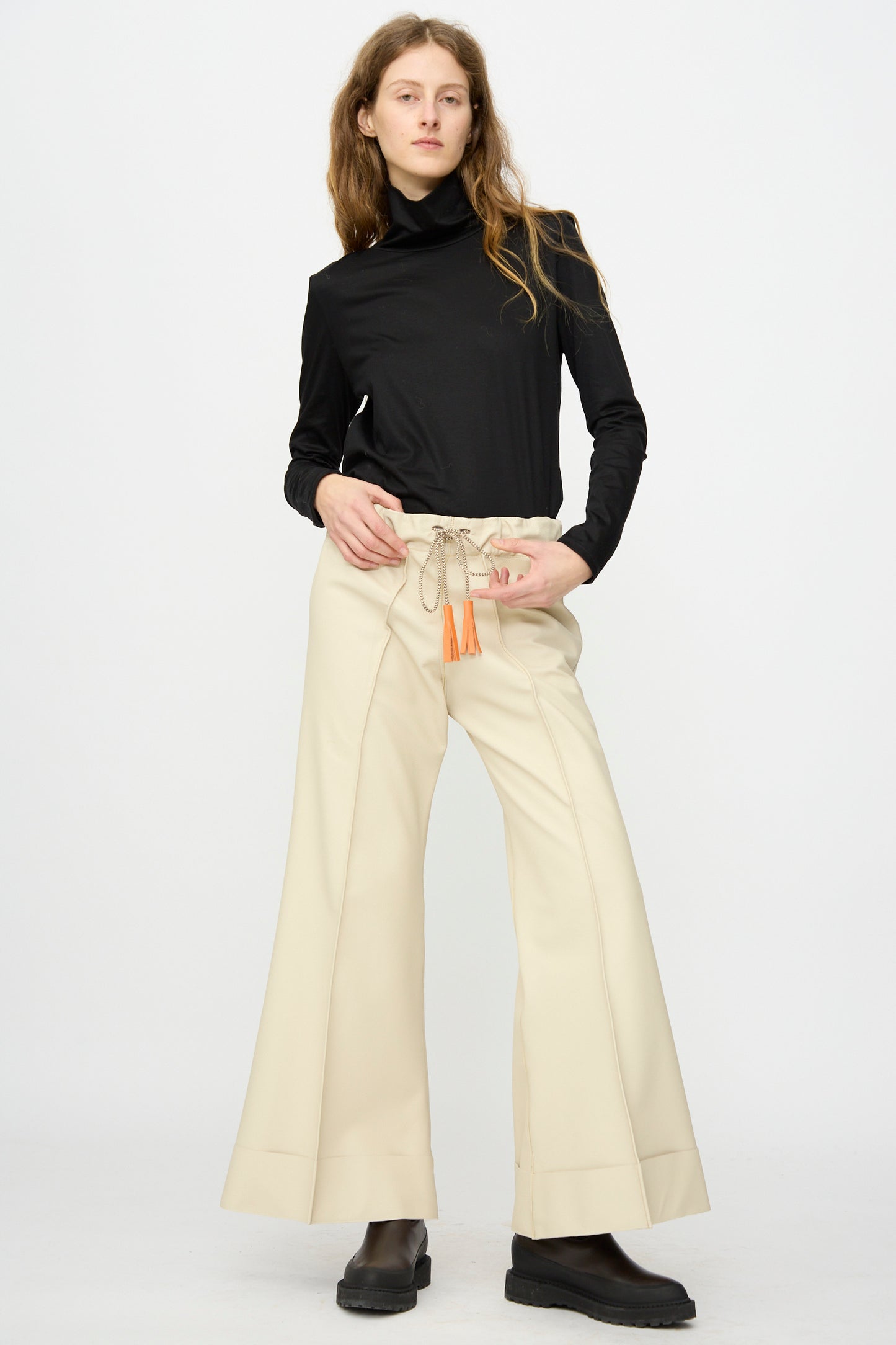 A person stands against a plain background wearing a black turtleneck, black boots, and Pierrot Flare Trouser in Leather by Thank You Have A Good Day.