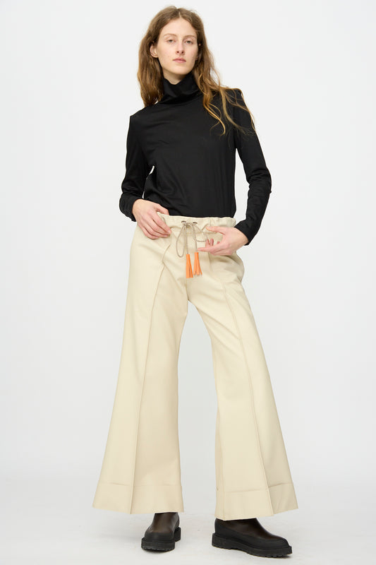 A person stands against a plain background wearing a black turtleneck, black boots, and Pierrot Flare Trouser in Leather by Thank You Have A Good Day.