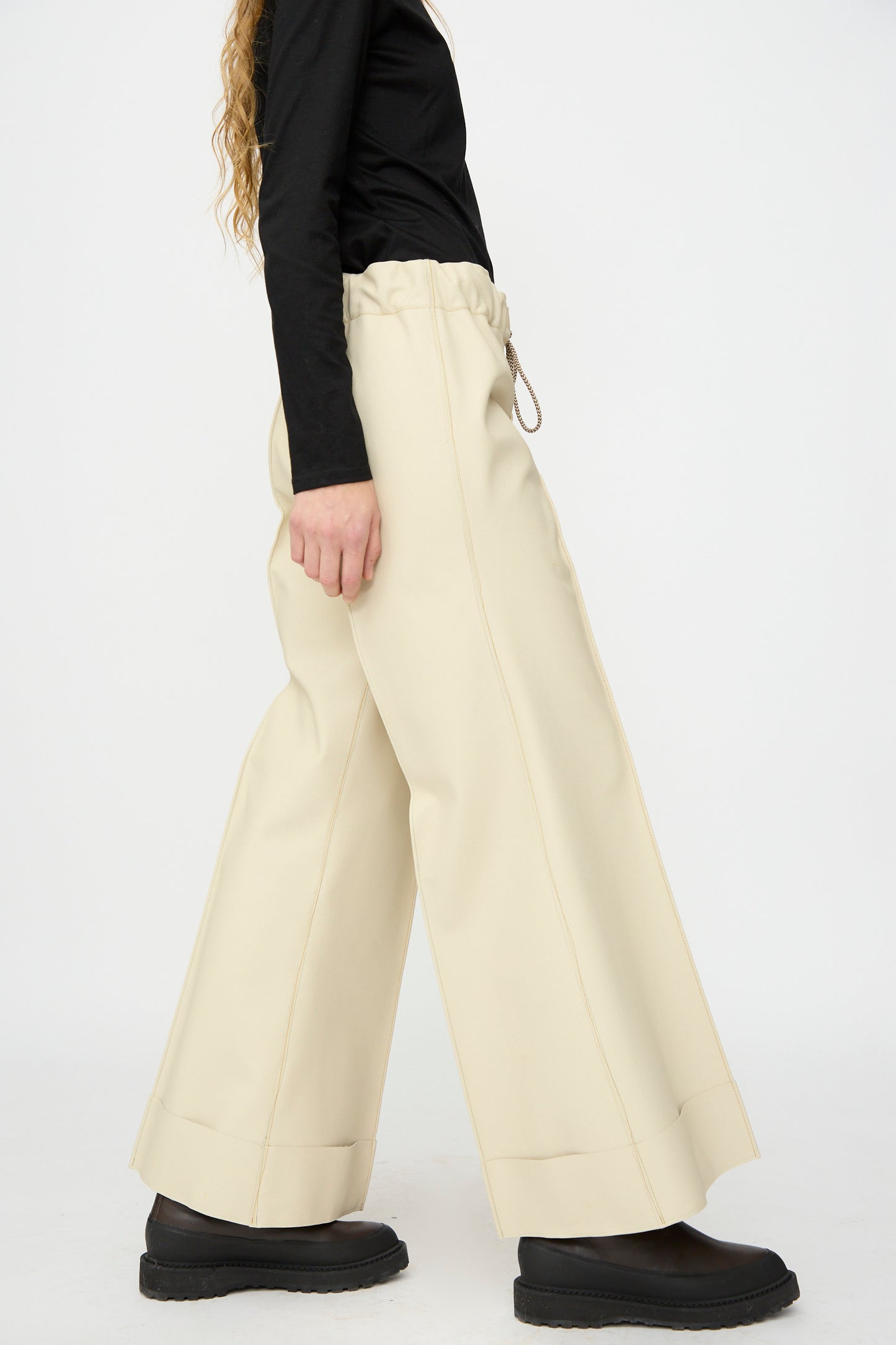 Person wearing a black long-sleeve top, Thank You Have A Good Day’s Pierrot Flare Trouser in Leather (cream, relaxed fit), and black platform shoes, standing against a plain white background.