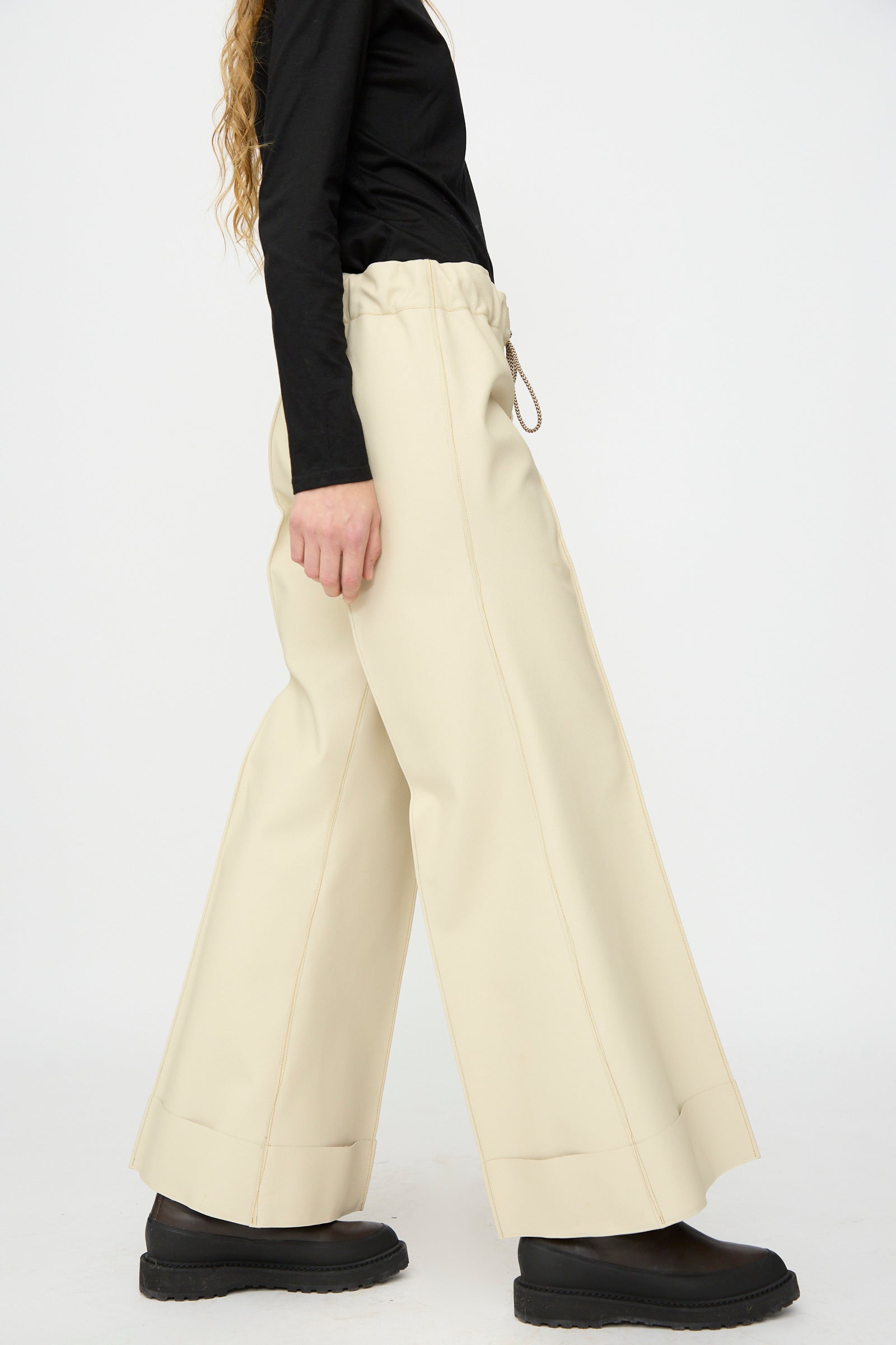 Person wearing a black long-sleeve top, Thank You Have A Good Day’s Pierrot Flare Trouser in Leather (cream, relaxed fit), and black platform shoes, standing against a plain white background.