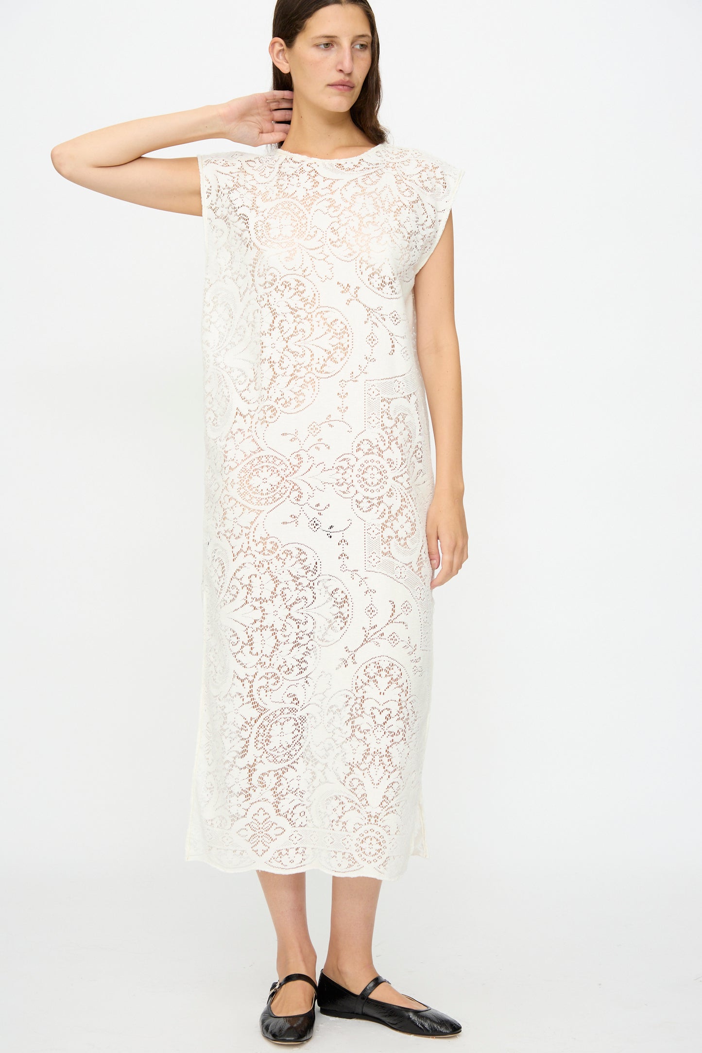 A woman stands against a white background wearing the Quaker Lace Caftan by Thank You Have A Good Day, an ankle-length, sleeveless white dress with a floral antique lace pattern, paired with black flat shoes.