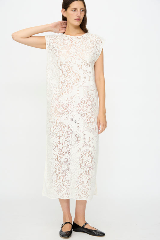 A woman stands against a white background wearing the Quaker Lace Caftan by Thank You Have A Good Day, an ankle-length, sleeveless white dress with a floral antique lace pattern, paired with black flat shoes.