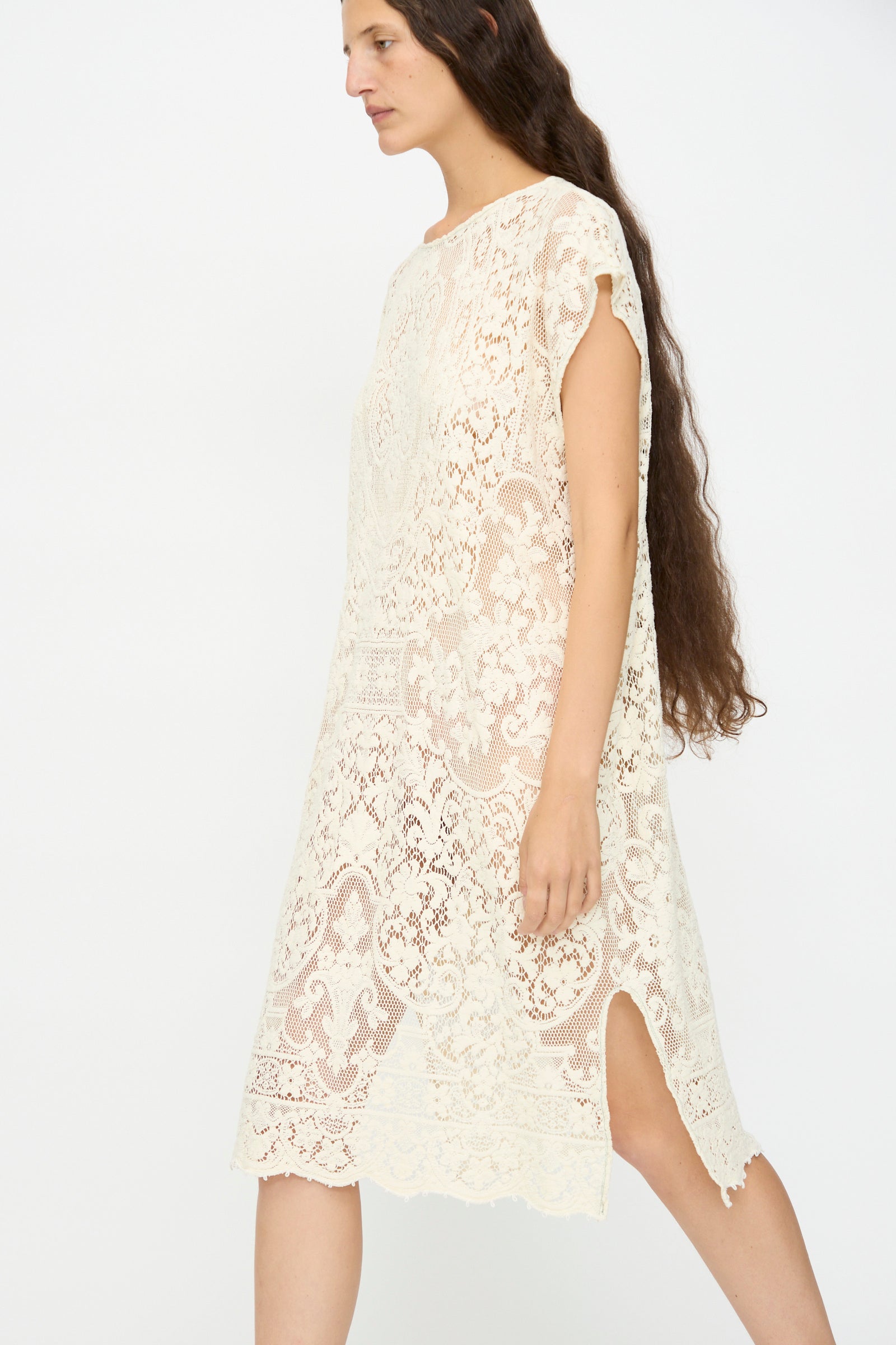 A woman with long brown hair wears the Thank You Have A Good Day Quaker Lace Caftan Midi Pullover—cream, loose-fitting, knee-length with short sleeves and side slit—walking against a plain white background.