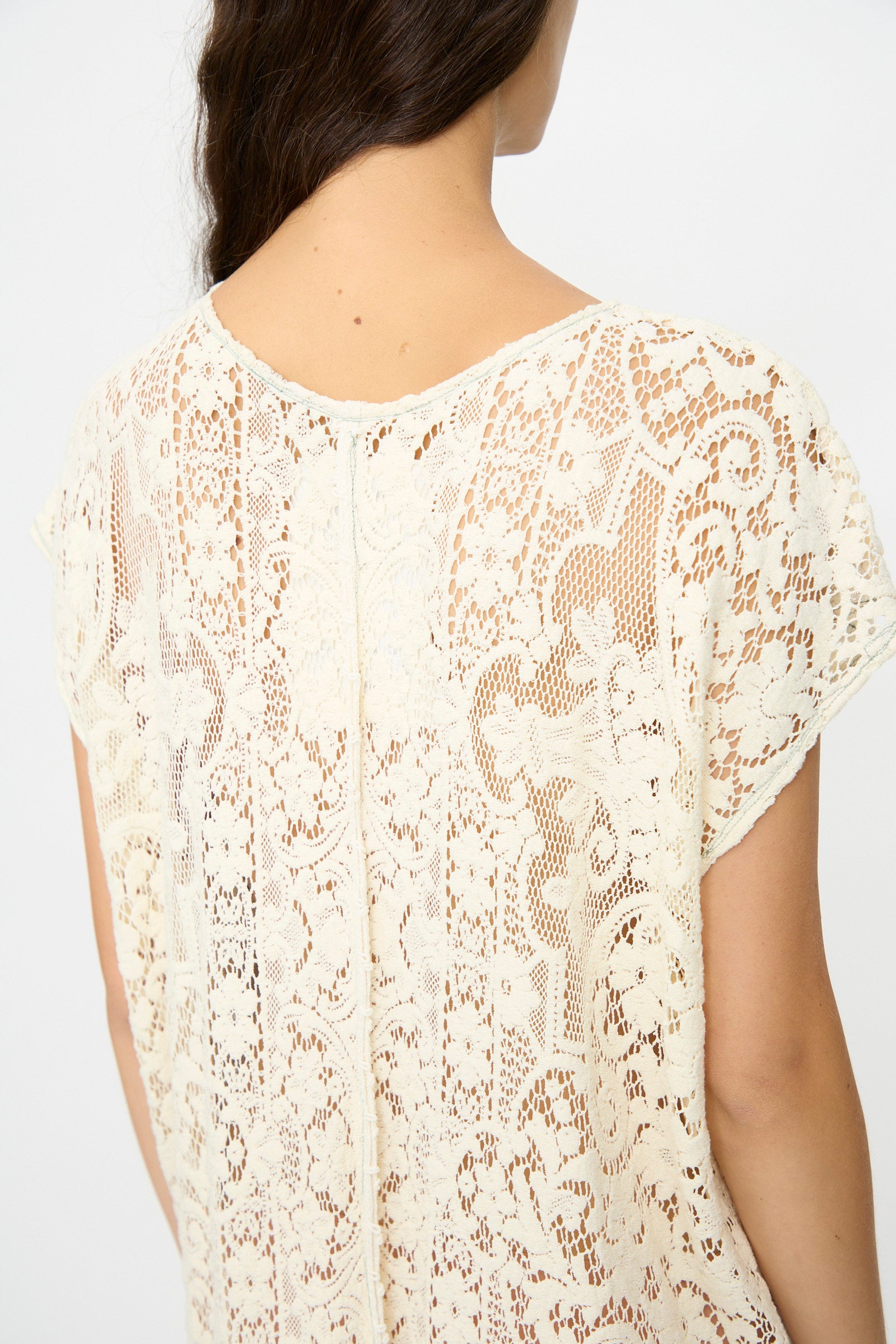 A person with long brown hair is seen from the back wearing the Quaker Lace Caftan Midi Pullover by Thank You Have A Good Day, an off-white, short-sleeve top featuring a detailed antique-inspired floral lace pattern.