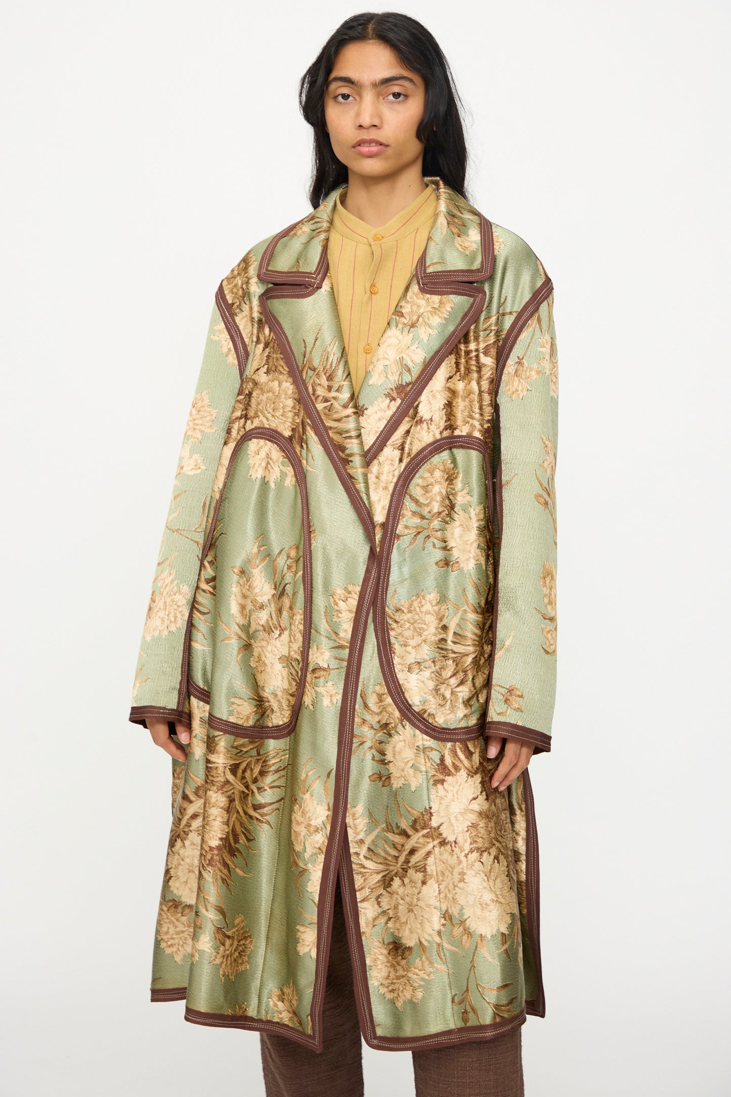 Someone wears the Thank You Have A Good Day Taped Trench in Floral—a handmade coat with a green floral pattern and brown trim—over a yellow shirt and brown pants, standing against a plain light background.