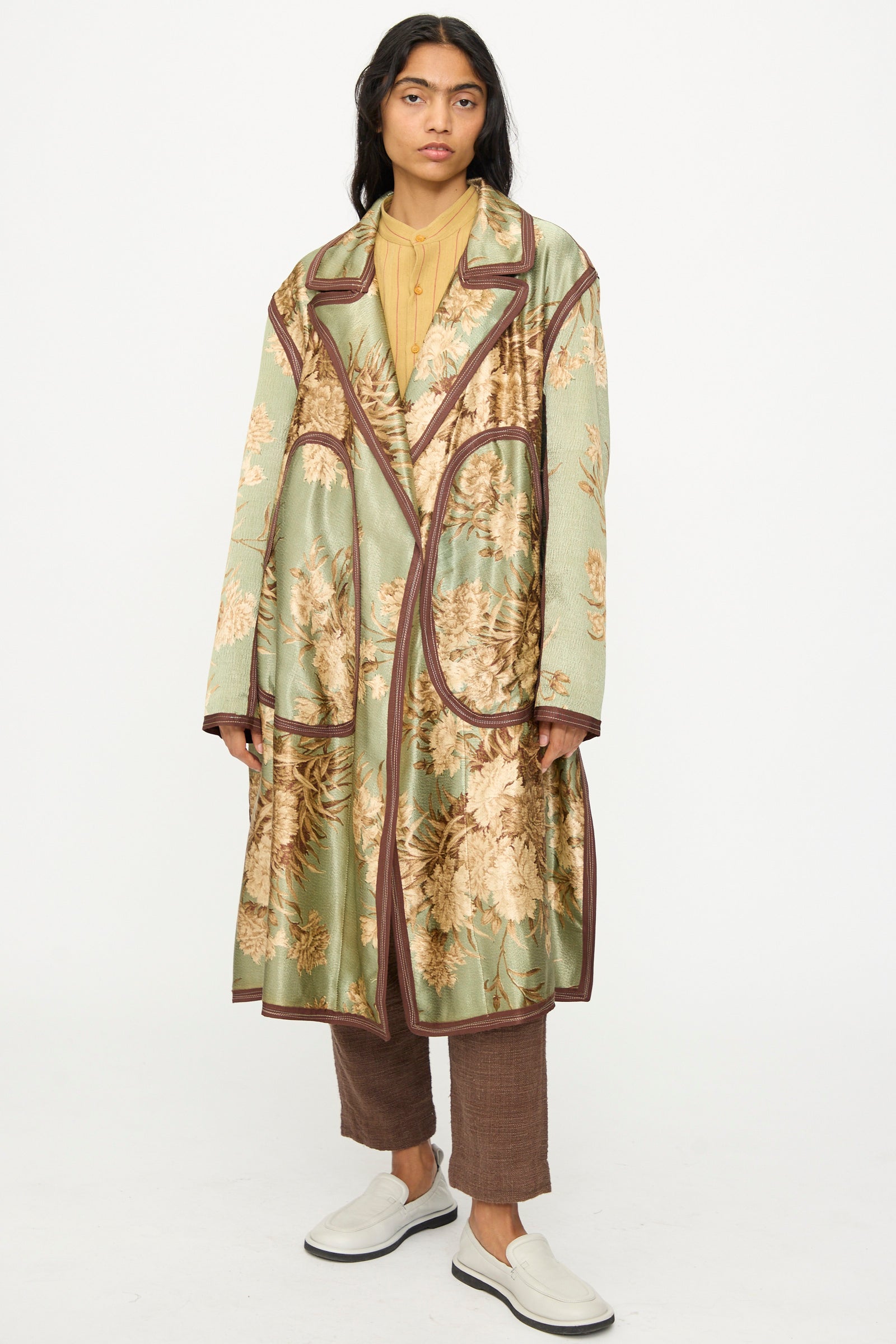 A person wears the Thank You Have A Good Day Taped Trench in Floral with a light brown shirt, brown pants, and white slip-on shoes, standing against a plain white background.