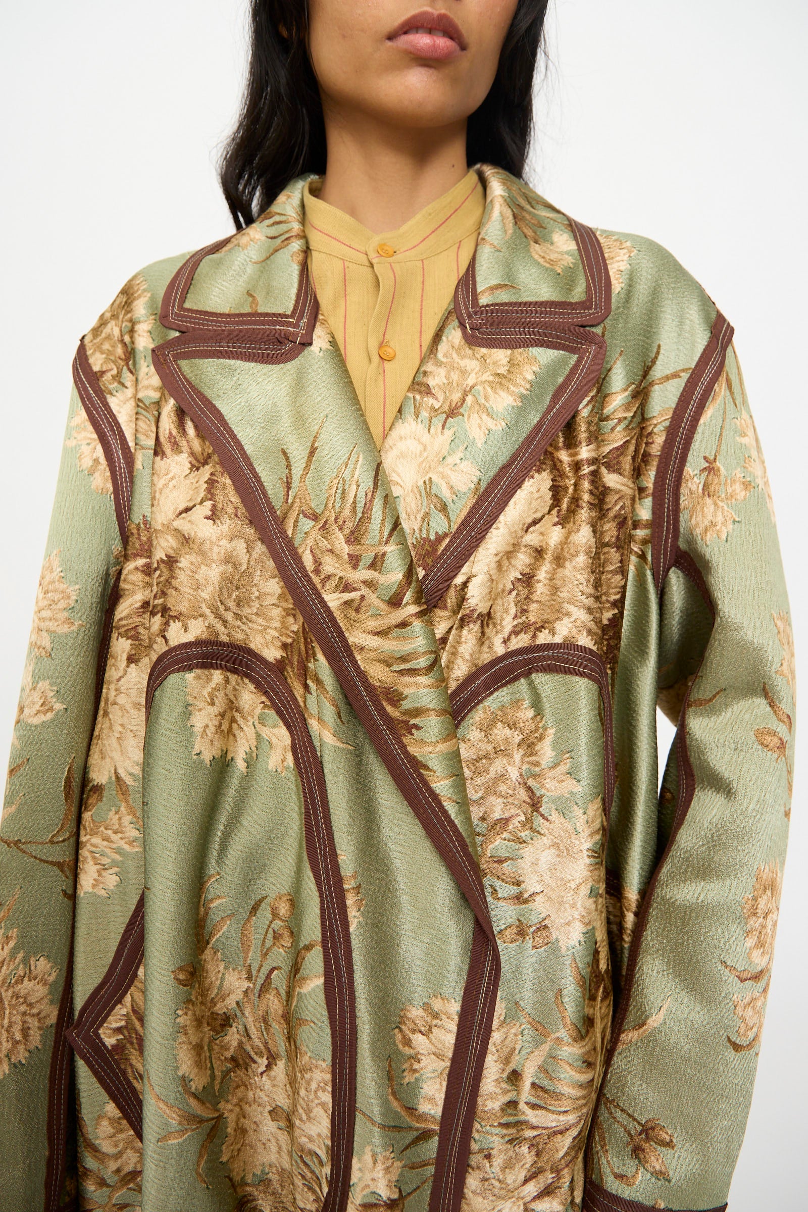 A person wears the Thank You Have A Good Day Taped Trench in Floral, featuring a green and beige vintage-inspired pattern with brown trim, layered over a yellow collared shirt, shown from shoulders to mid-torso.