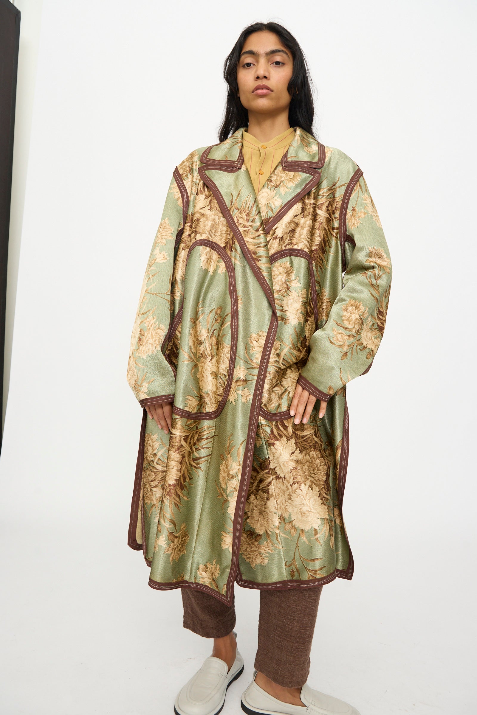 A person stands against a plain white background, wearing the Thank You Have A Good Day Taped Trench in Floral with brown trim, brown pants, and white loafers.